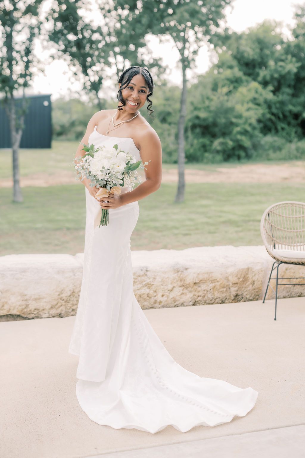 Elegant bride in white fitted gown holding white floral bouquet smiling outdoors on wedding day.