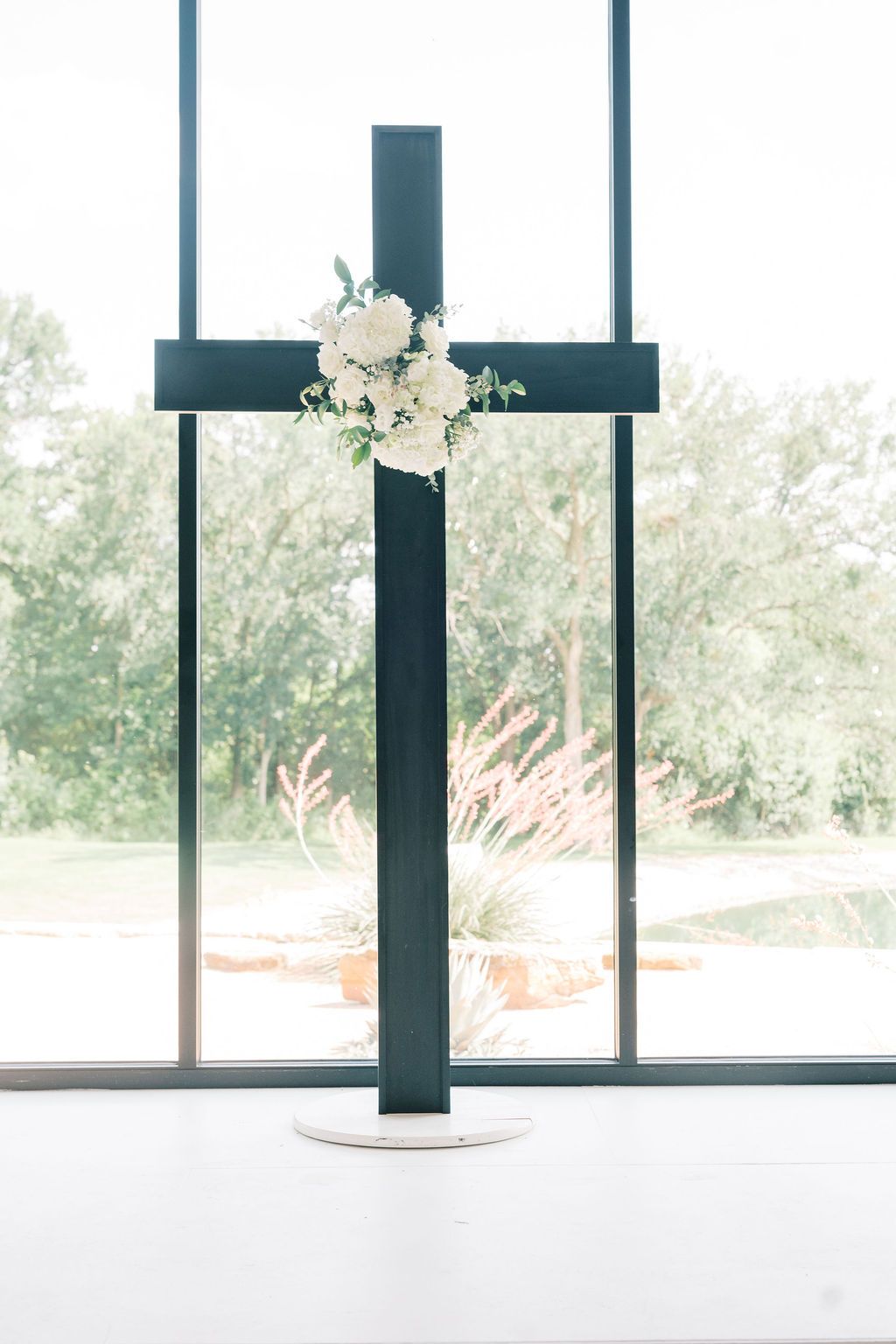 Dark cross decorated with white roses and greenery stands before a large window at a wedding ceremony.