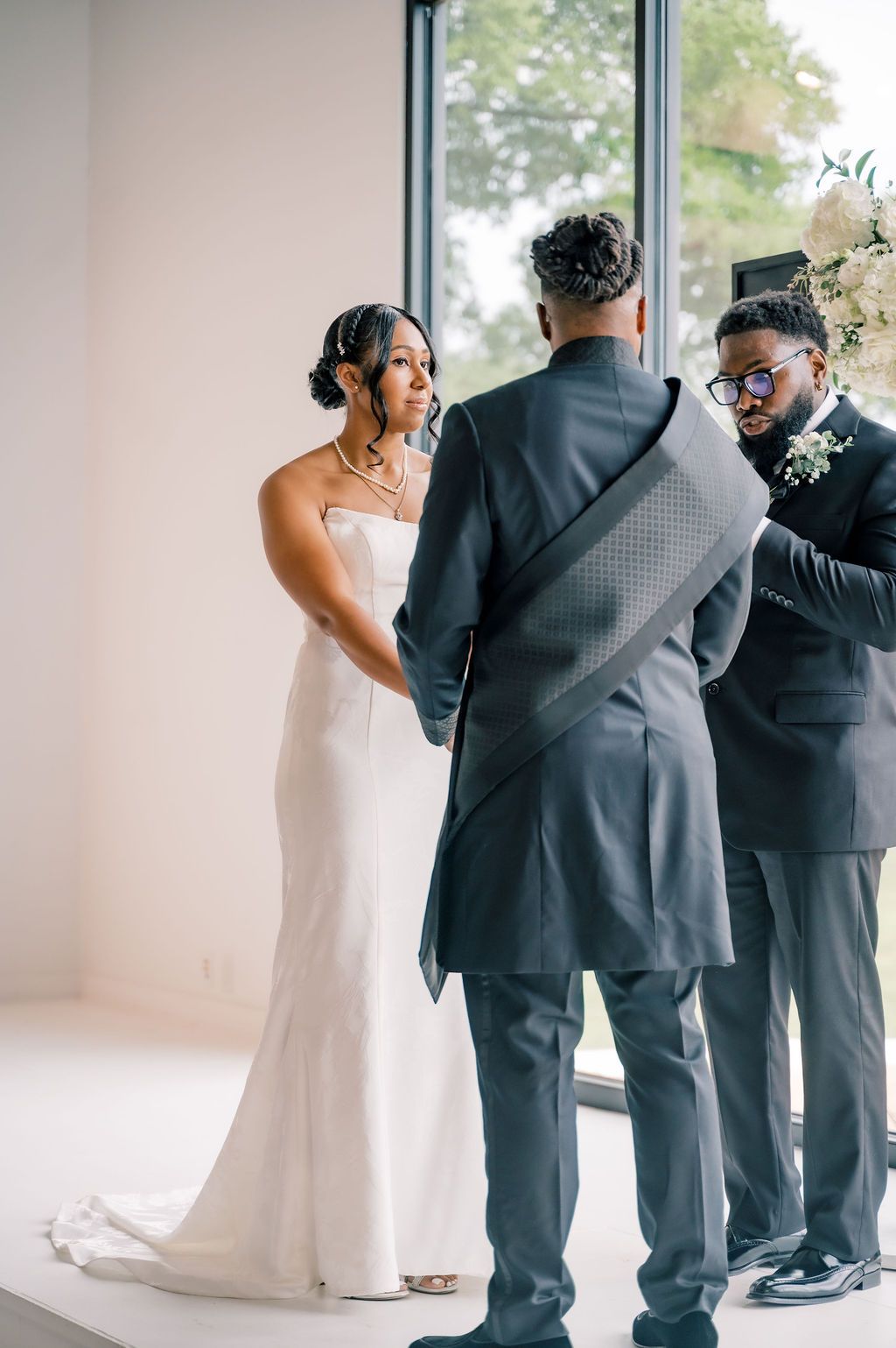 Bride in white gown holds hands with groom in gray suit during wedding ceremony indoors.
