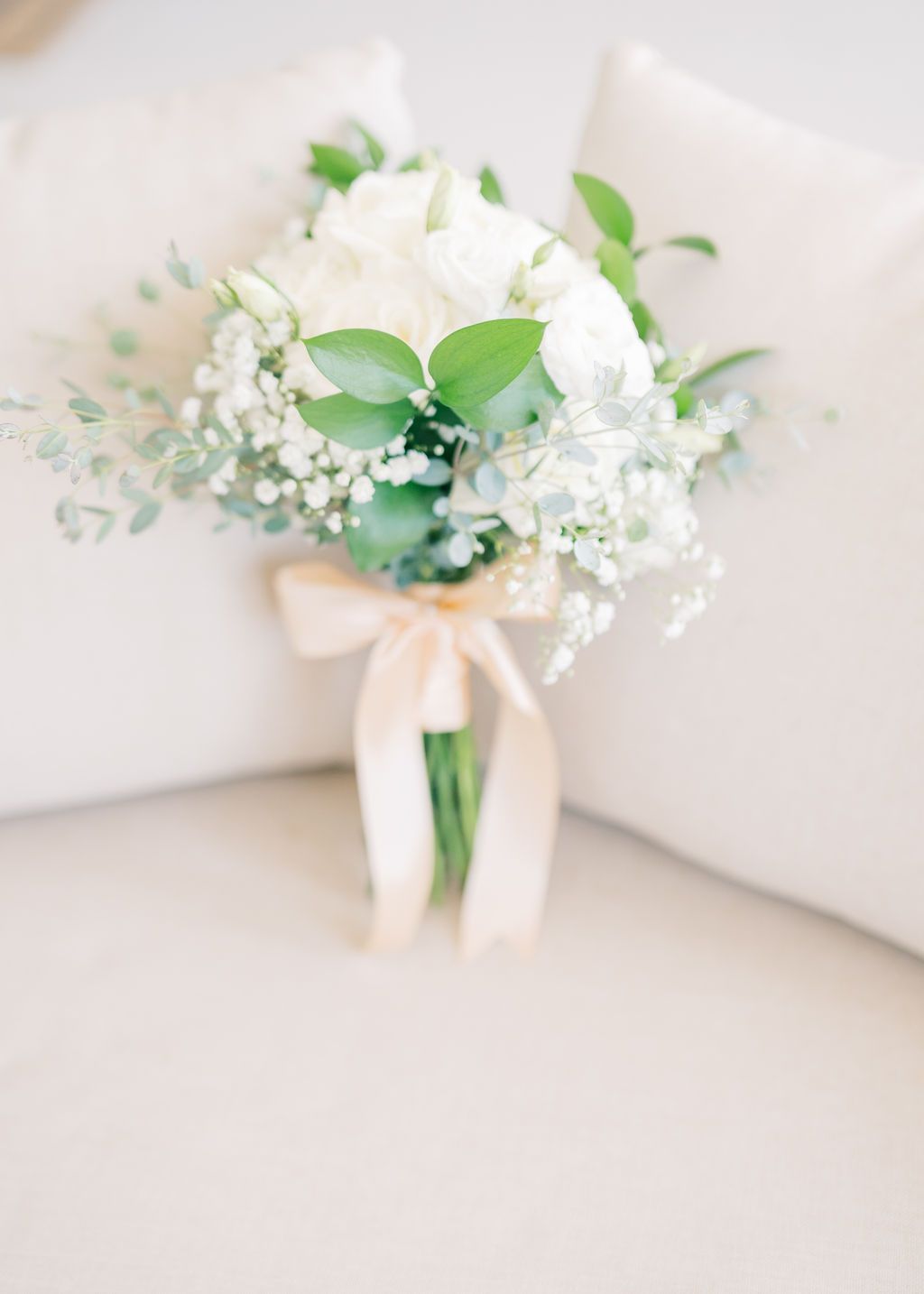 Elegant bridal bouquet of white roses, baby's breath, and greenery tied with a blush satin ribbon on a white chair.
