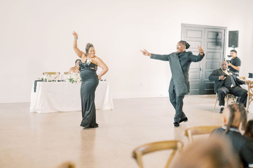Couple dancing energetically at wedding reception in elegant white venue, wearing formal black attire.