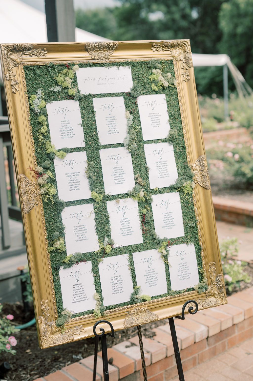 Gold ornate framed wedding seating chart with moss background and white cards displayed on a black easel outdoors.