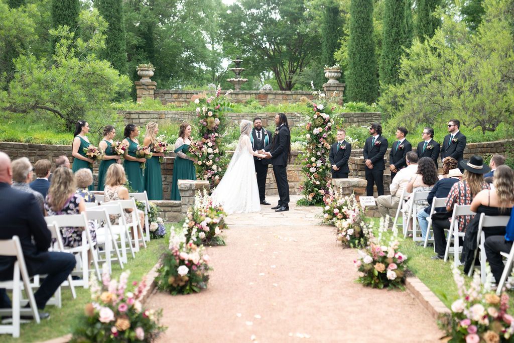 Outdoor wedding ceremony with bride and groom at floral arch, bridesmaids in teal, groomsmen in black, white chairs, garden venue.