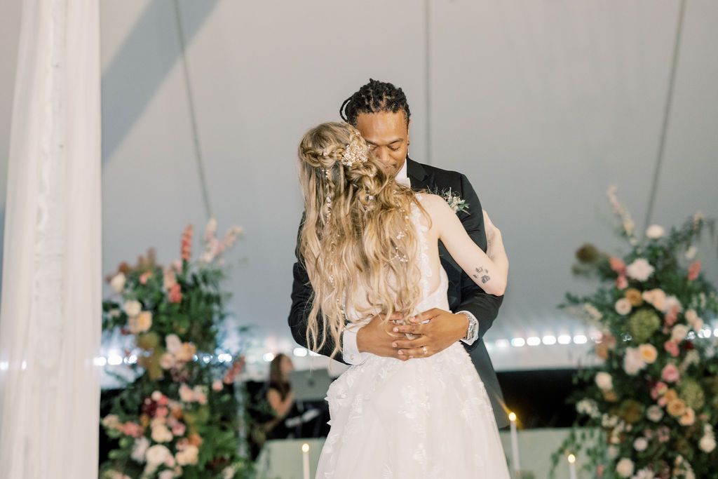 Bride and groom share first dance under white tent surrounded by lush floral arrangements at elegant wedding reception.