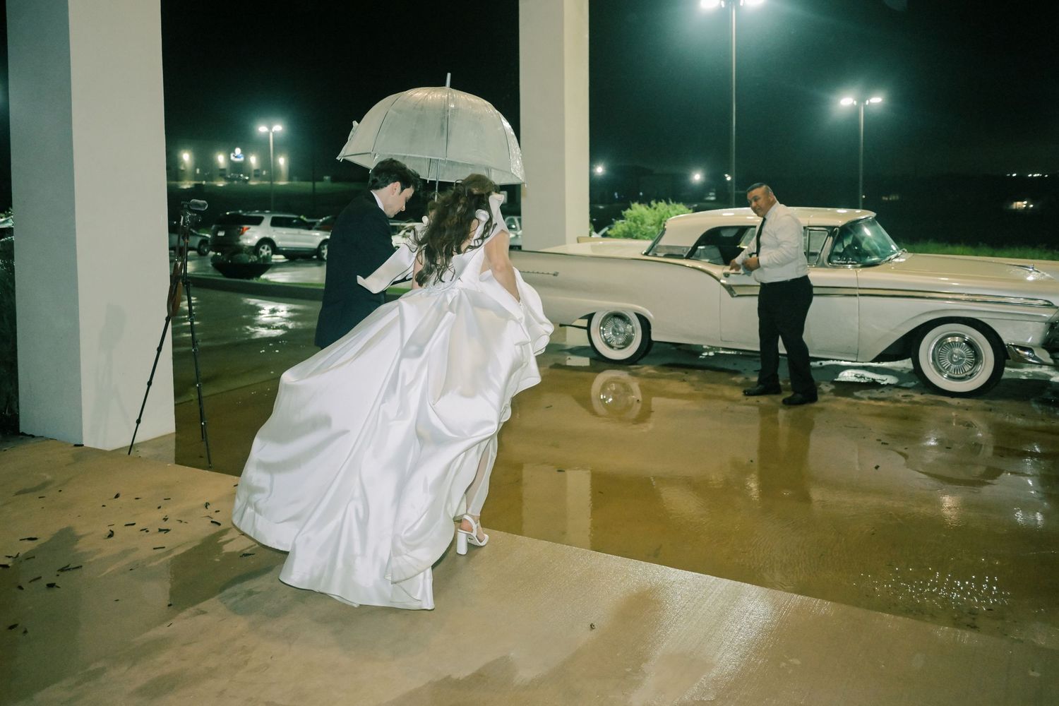 Bride and groom walking under umbrella in rain beside vintage classic car at night.