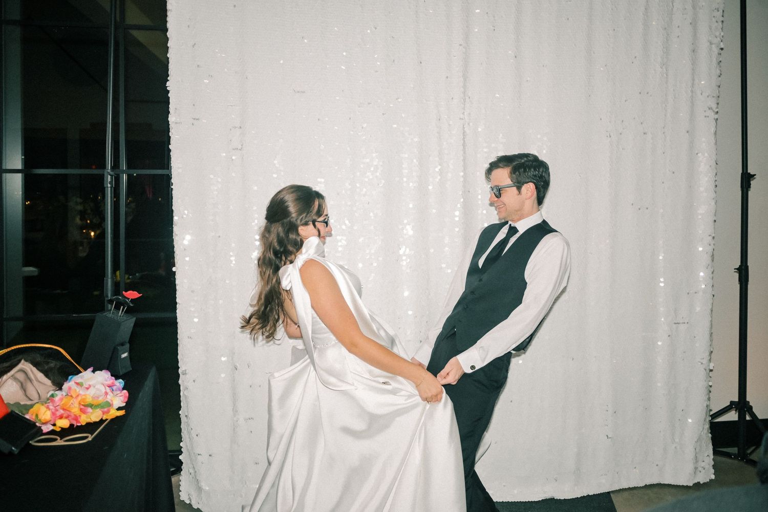 Bride and groom holding hands in front of white sequin backdrop at wedding reception.