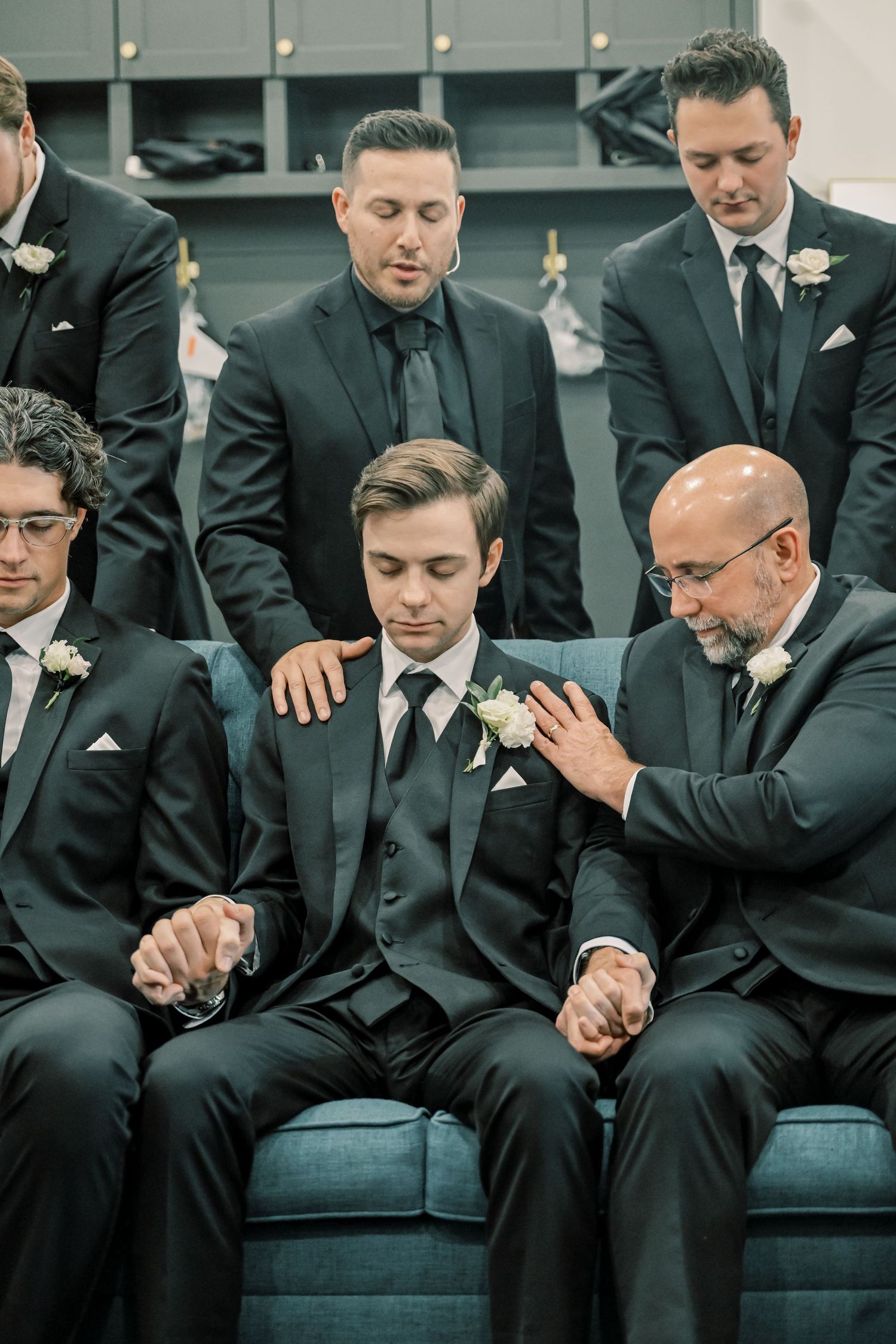 Groomsmen in black suits gather around the groom for a prayer moment before the wedding ceremony.