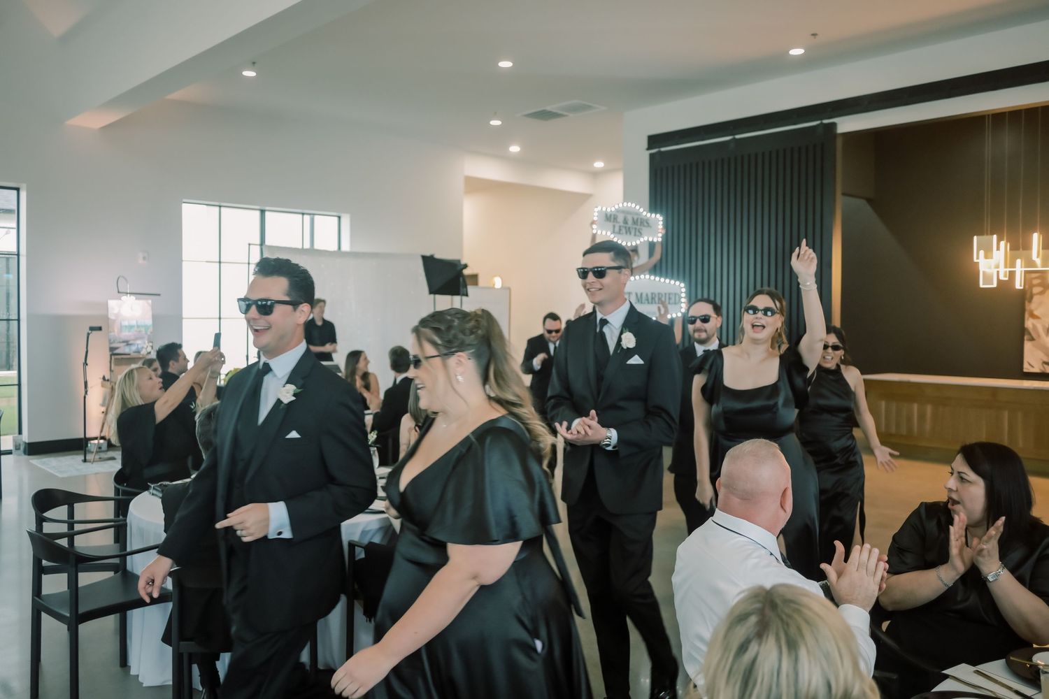 Wedding party in black attire wearing sunglasses making grand entrance at reception celebration.