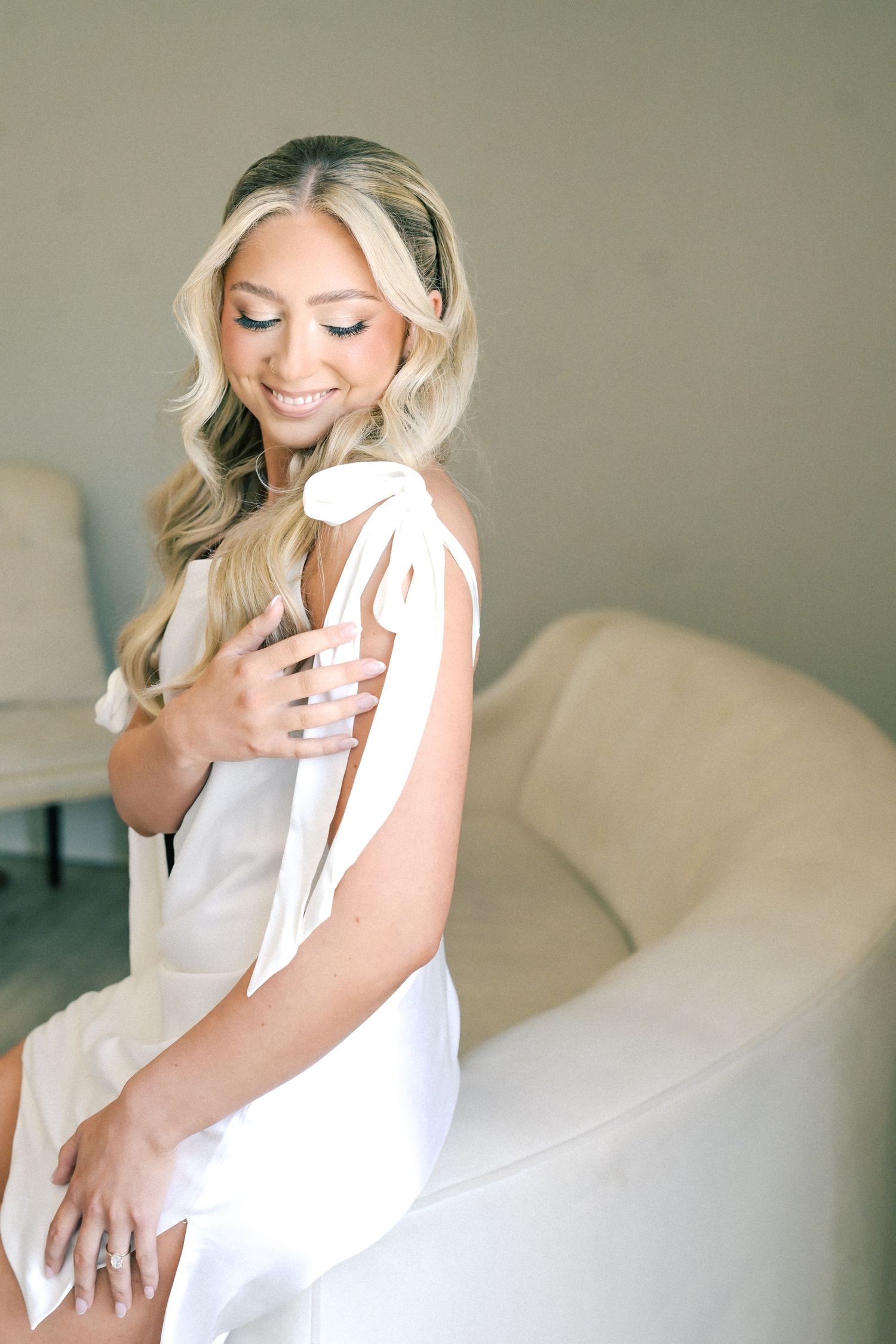 Smiling blonde bride in white satin bow-shoulder dress sitting on cream sofa with engagement ring.