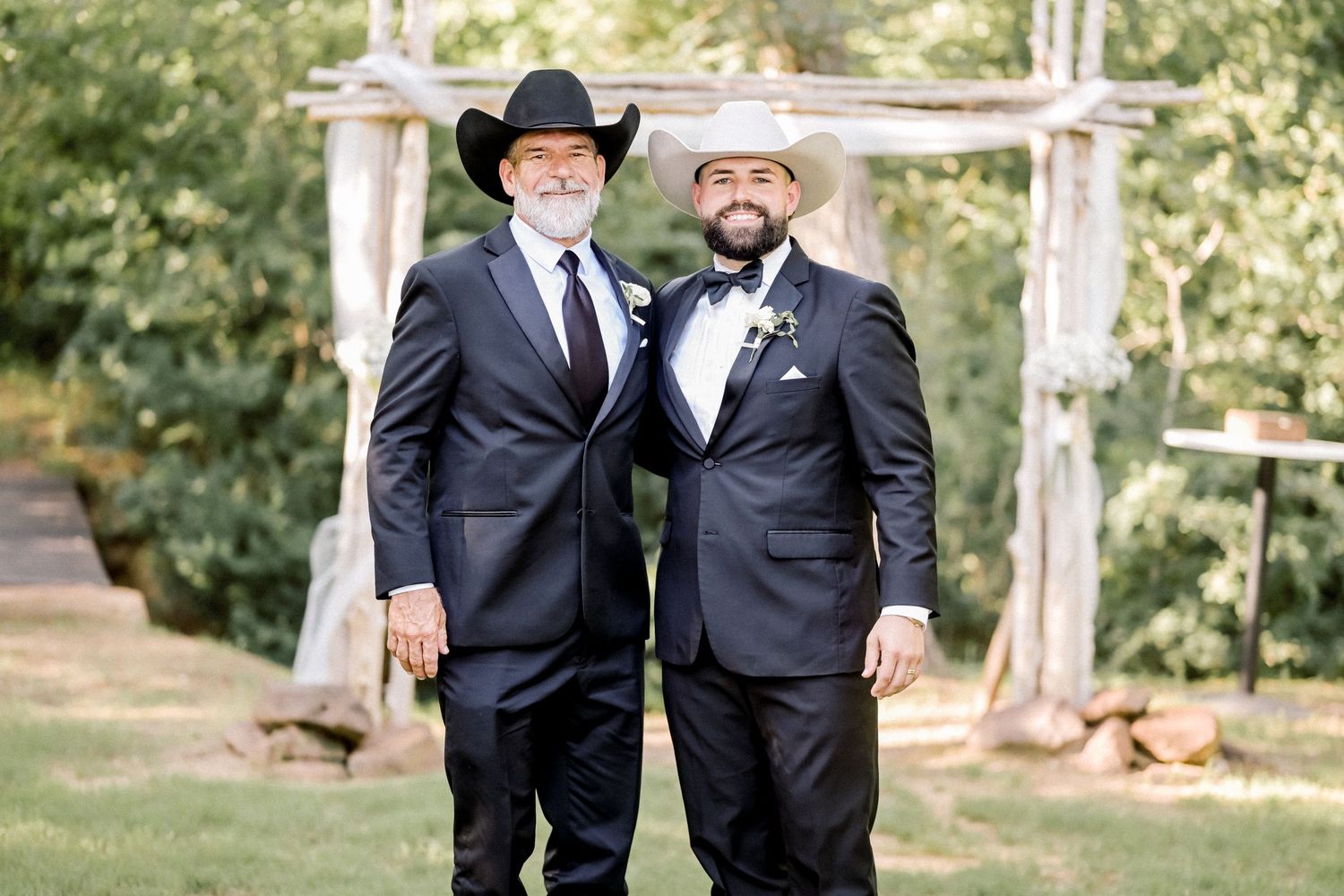 Two men in dark suits and cowboy hats posing together outdoors near a rustic wooden arch.