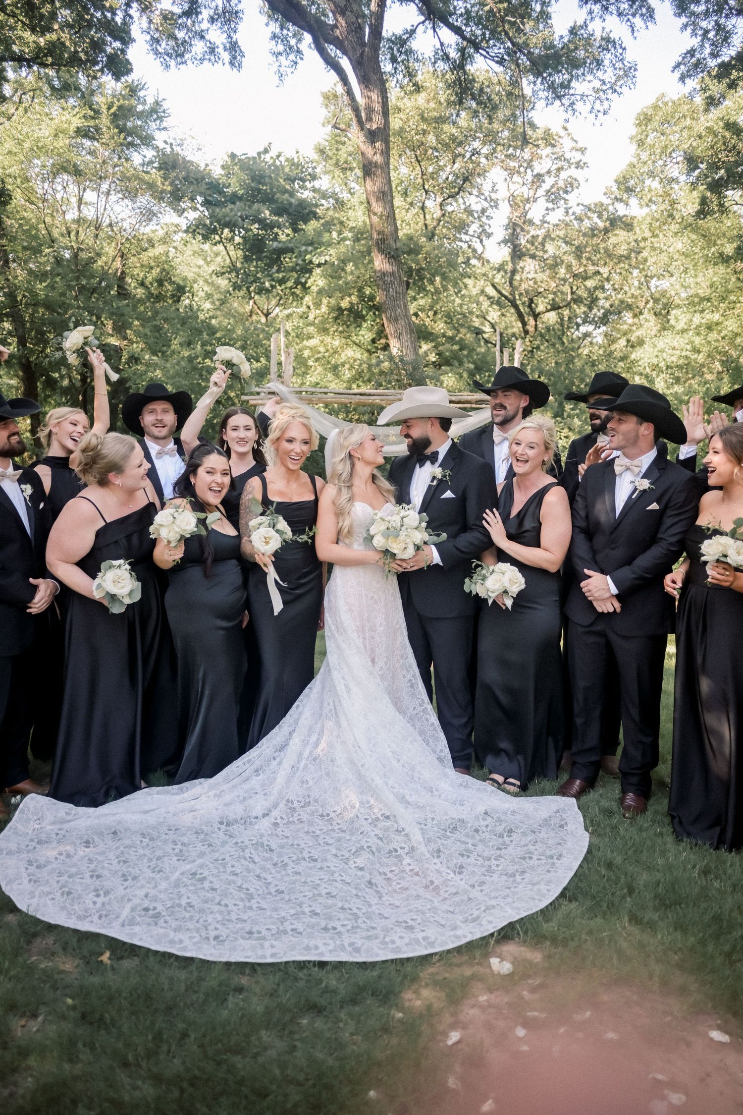 Joyful wedding party celebrates with bride and groom in lace dress outdoors under trees.