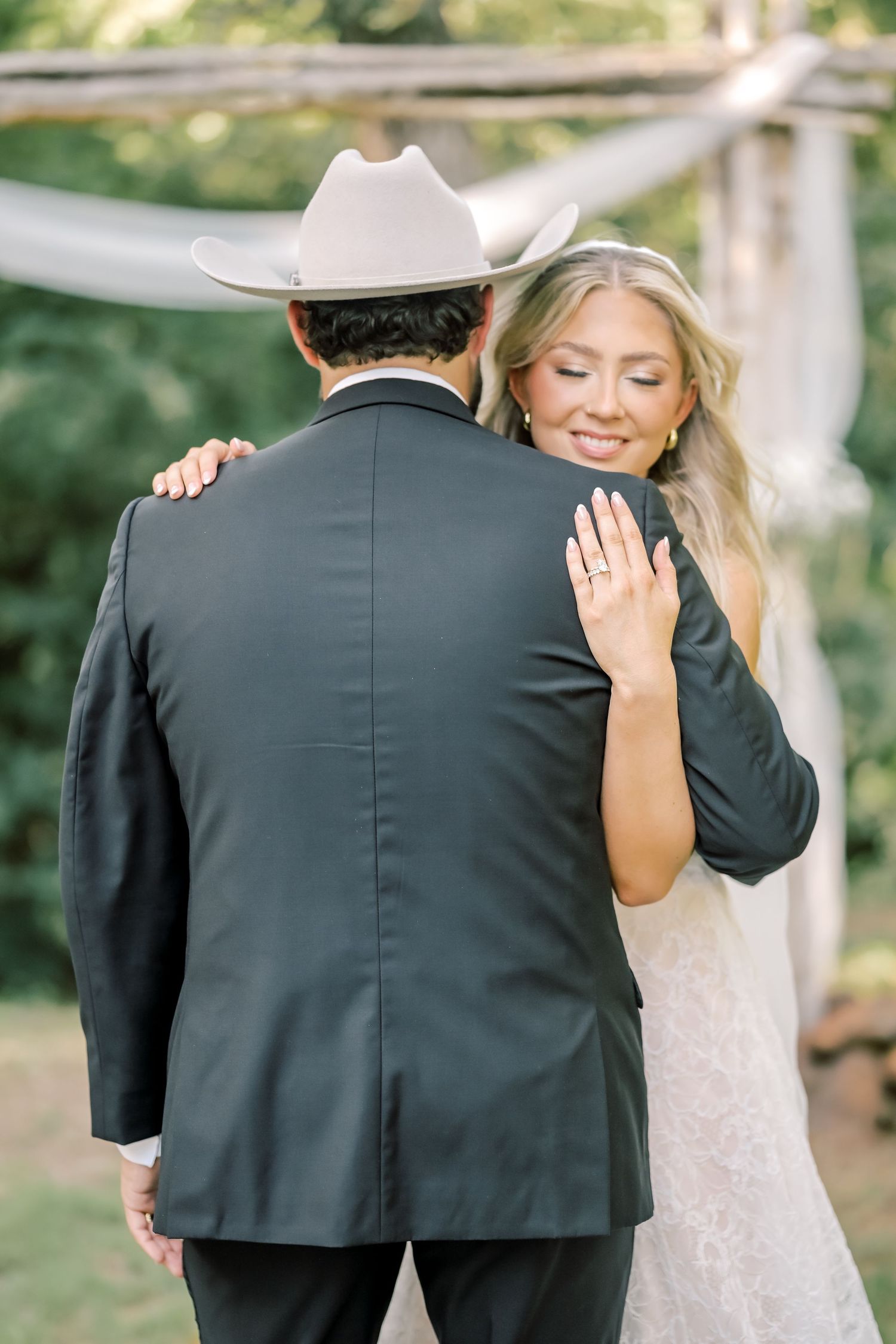 Bride in lace wedding gown embracing groom in black tuxedo and white cowboy hat at ceremony.