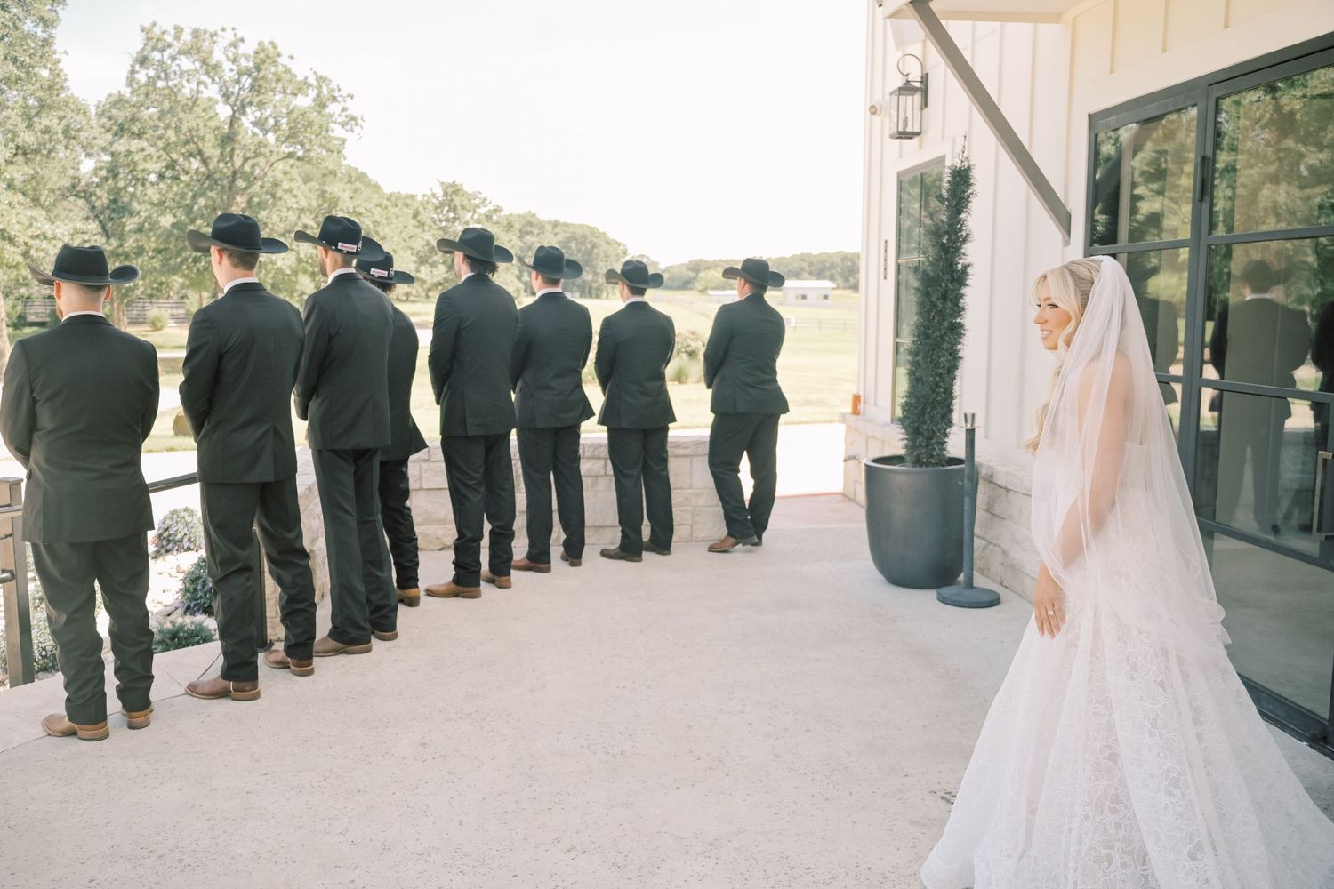 Bride in lace gown and veil stands near groomsmen in black cowboy hats outside a barn venue.