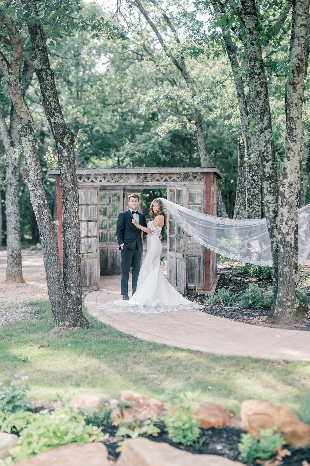 Bride and groom stand together outdoors among tall trees, her veil flowing in the wind near a rustic wooden arbor.