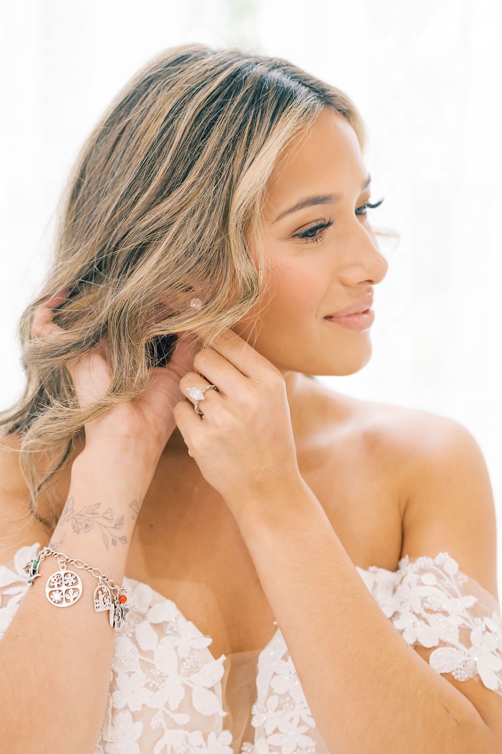 Bride in lace gown puts on earring, showcasing engagement ring and charm bracelet with floral tattoo.