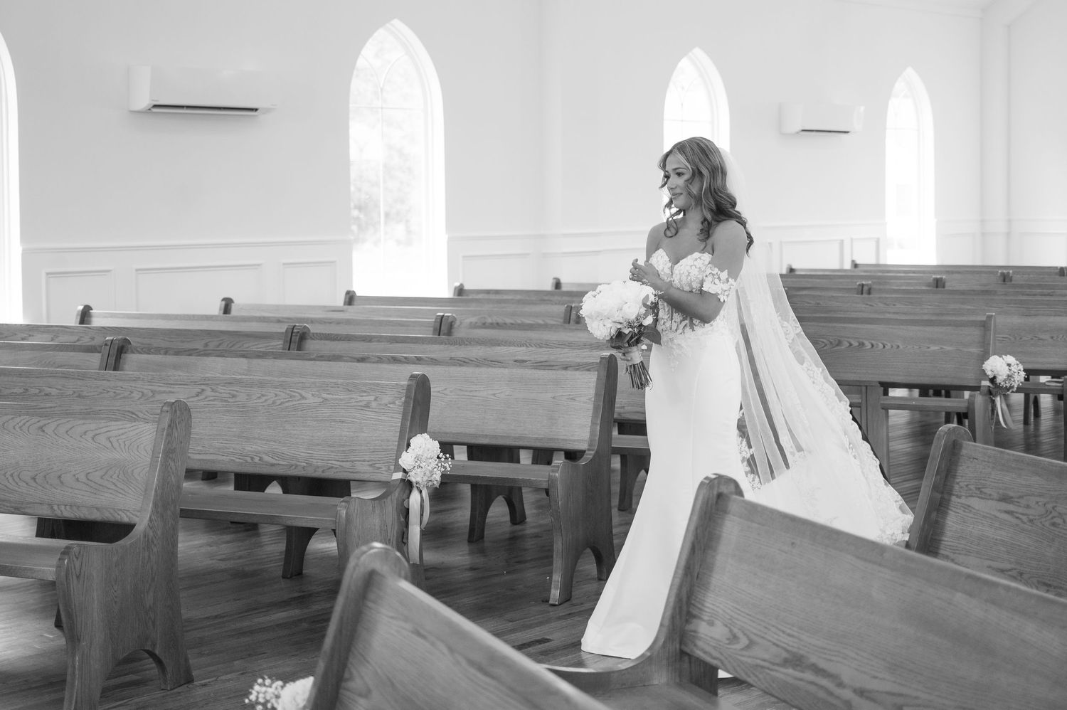 Bride in elegant white gown walks down church aisle holding bouquet in black and white photo.