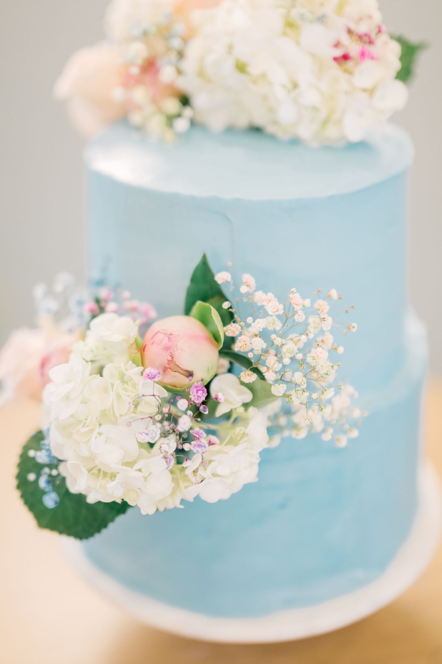 Two-tier light blue wedding cake decorated with white hydrangeas, pink peonies, and baby's breath flowers.