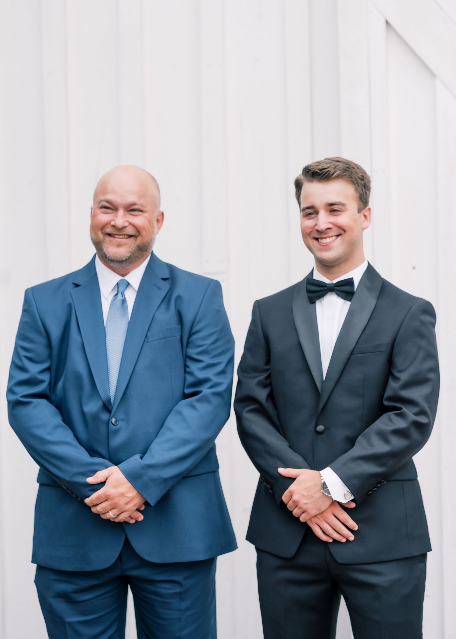 Groom in black tuxedo posing with man in blue suit smiling in front of white barn wall.