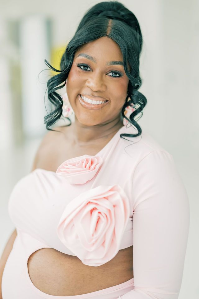 Smiling Black woman in pink one-shoulder top with rose detail, elegant updo hairstyle, posing confidently indoors.