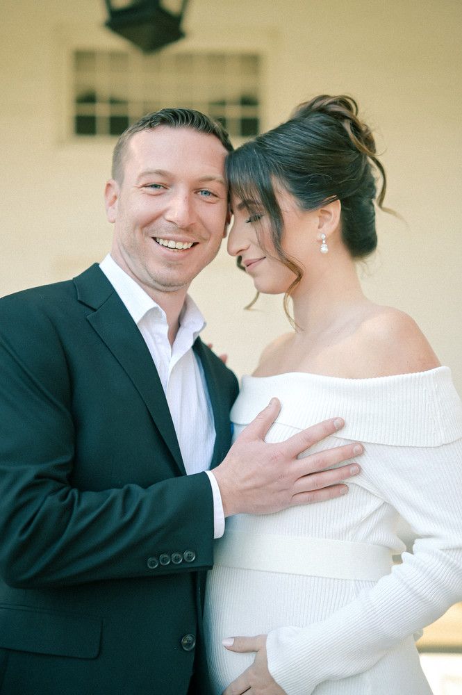 Smiling couple in elegant attire pose for maternity photo, man in dark suit embracing pregnant woman in white off-shoulder top.