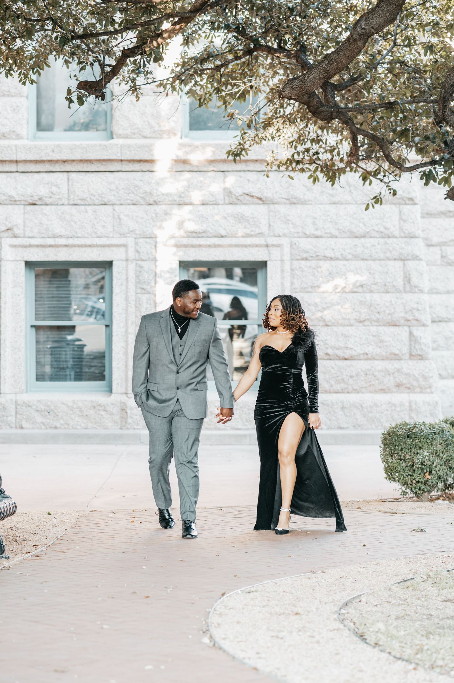 Elegant couple walking hand-in-hand, man in gray suit and woman in black velvet gown, outside a stone building.