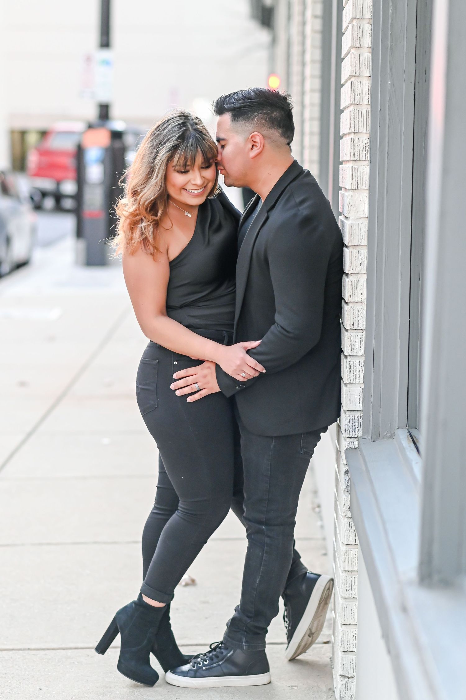 Couple in black outfits sharing a tender moment leaning against a building on a city street.