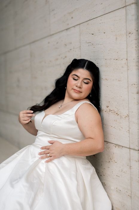 Bride in a white satin gown leaning gracefully against a stone wall, eyes closed, outdoors.