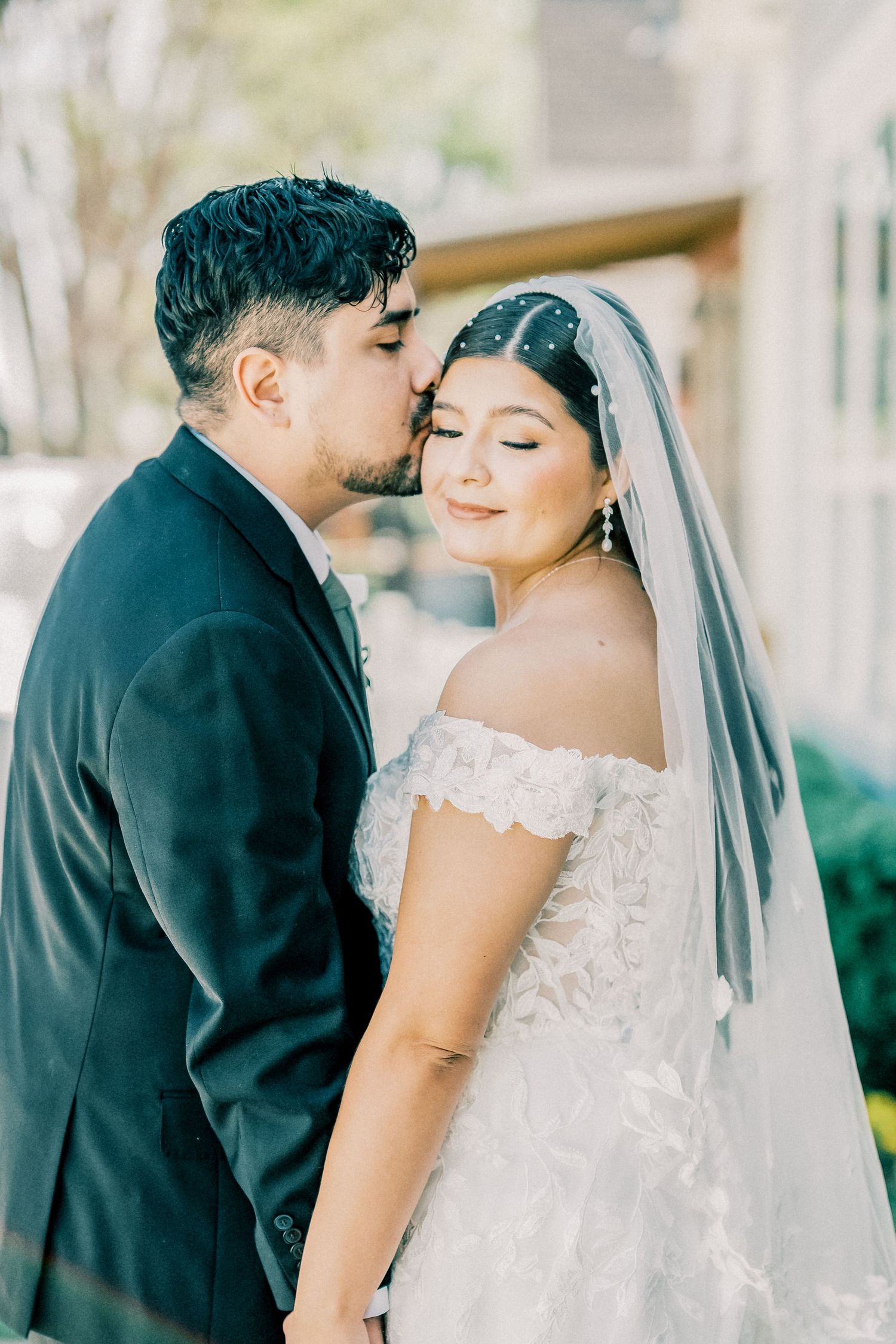 Groom in dark suit kissing bride in off-shoulder lace gown and veil on the cheek during outdoor wedding portrait.