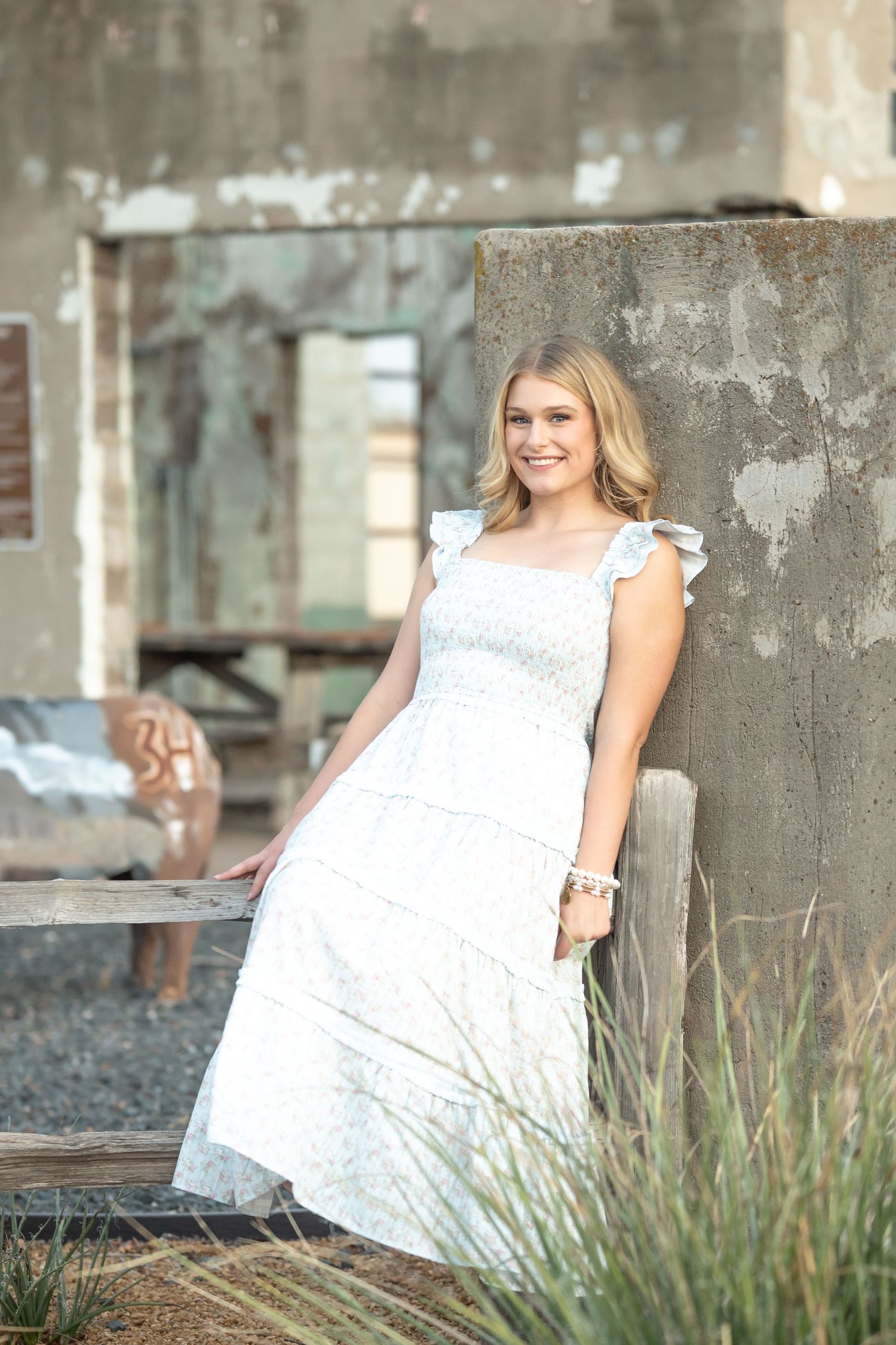 Young woman in white floral midi dress leaning against concrete wall outdoors, smiling warmly in natural light.
