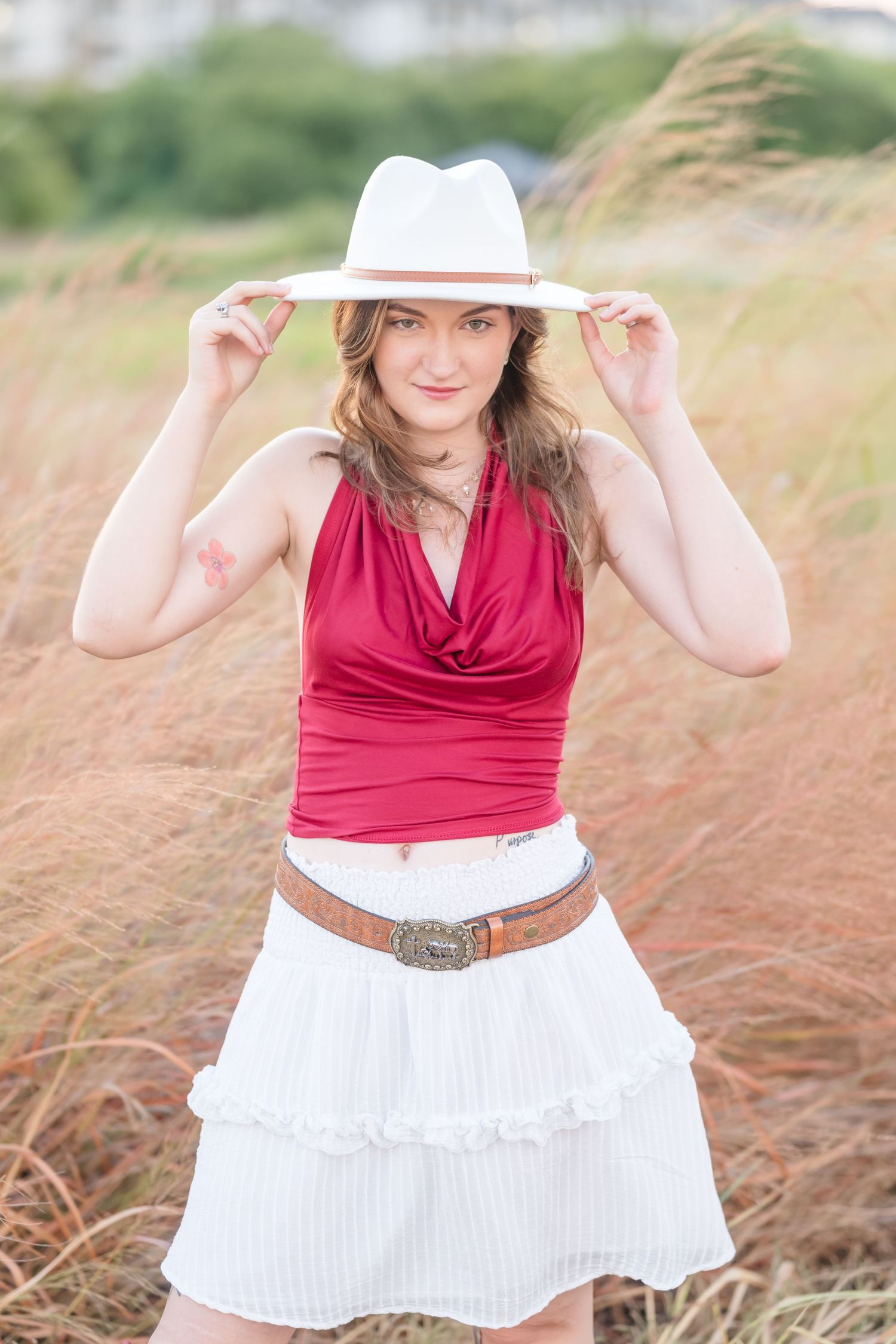 Young woman in red top, white ruffled skirt, and white fedora hat posing in a golden grass field.