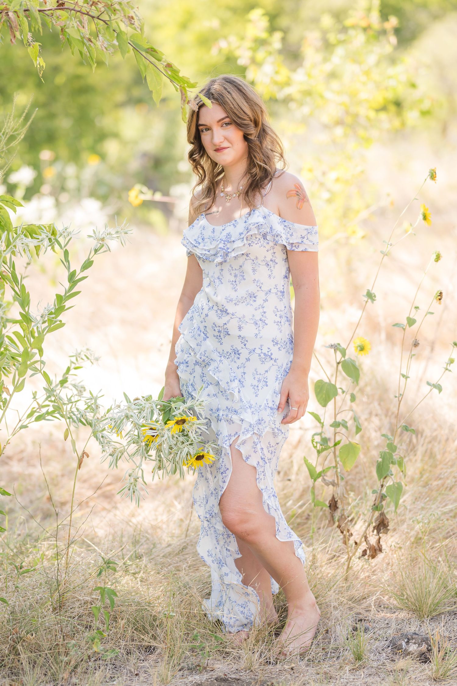 Young woman in blue floral maxi dress holding sunflowers among wildflowers outdoors.