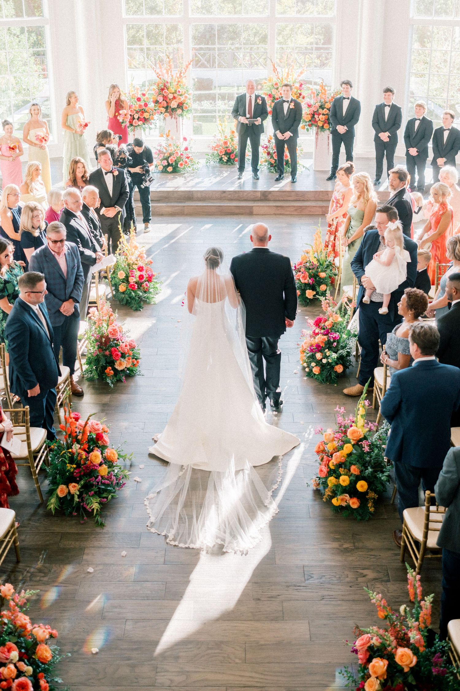 Wedding ceremony in a bright venue with orange and pink floral aisle decorations.