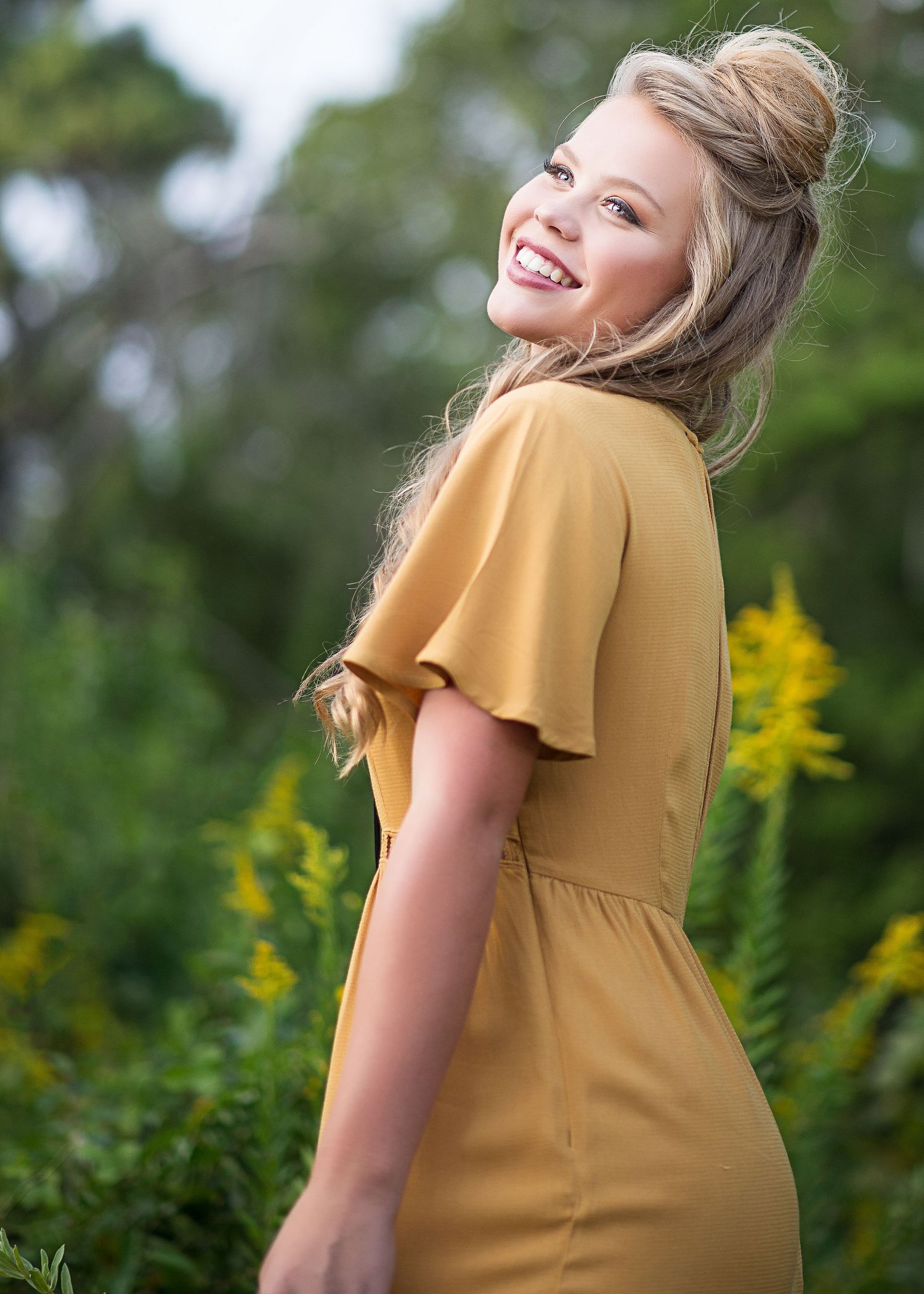 A joyful person wearing a mustard yellow dress poses outdoors with yellow wildflowers and greenery in the background.