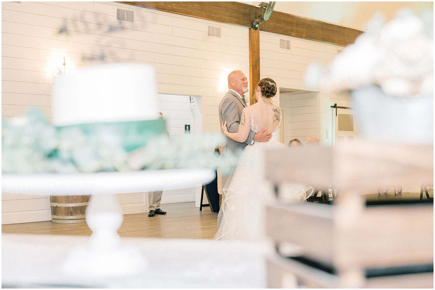 A couple dances together in an elegant white reception hall with exposed wooden beams overhead.