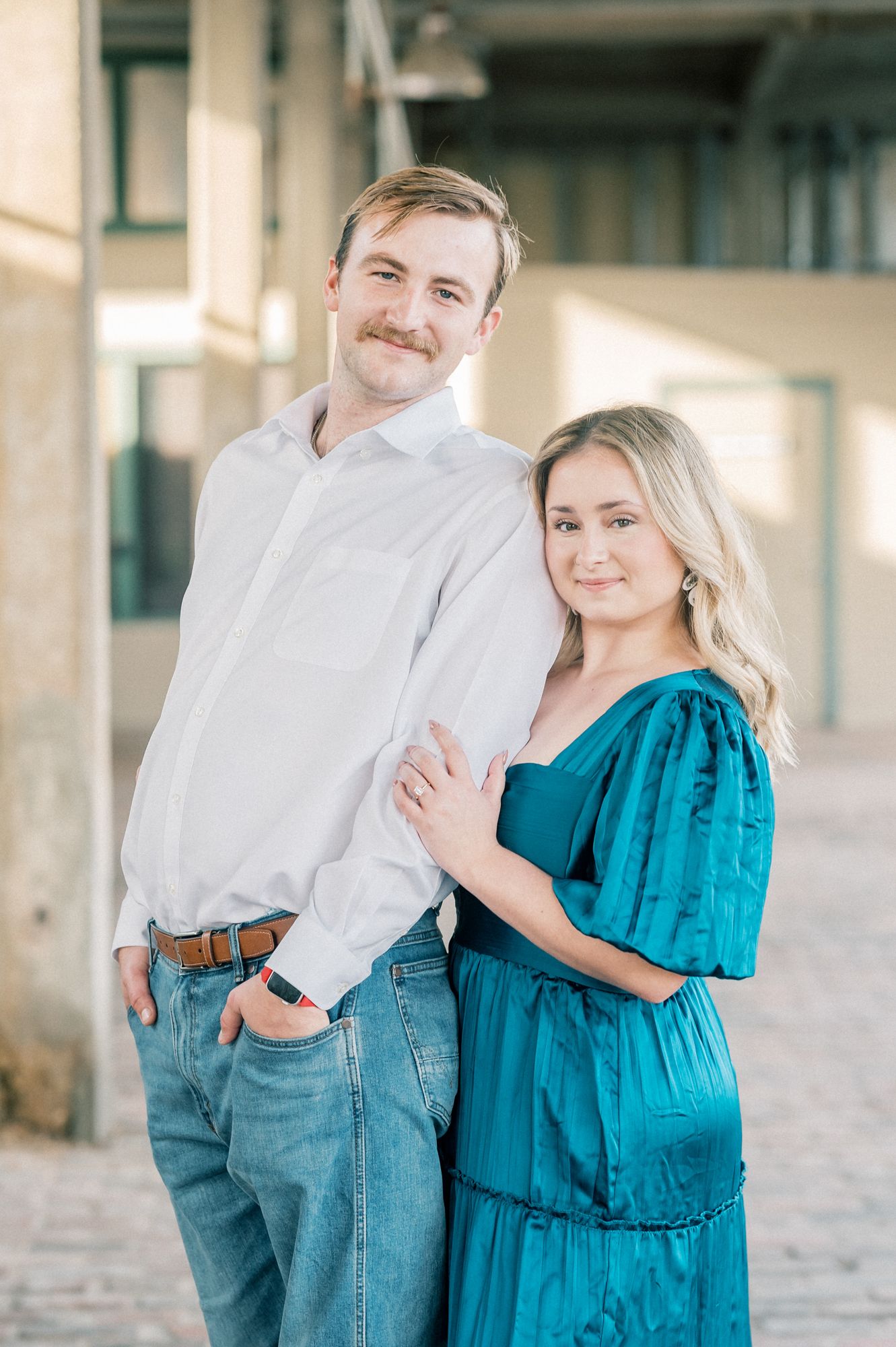 A couple embraces in a bright indoor space with columns, wearing white and turquoise blue outfits.