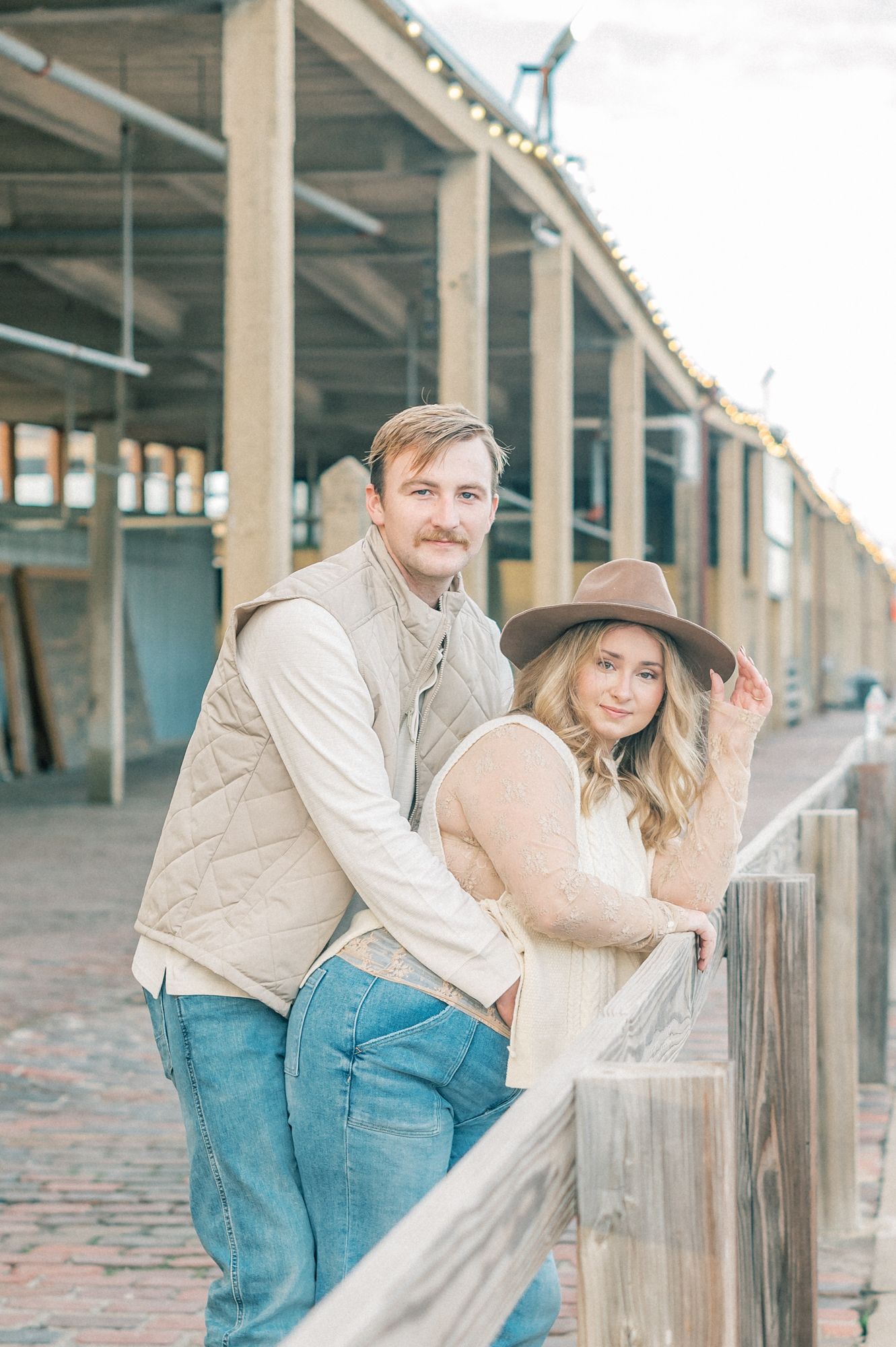 A couple in coordinating beige outfits poses casually against a wooden railing at a rustic outdoor location.