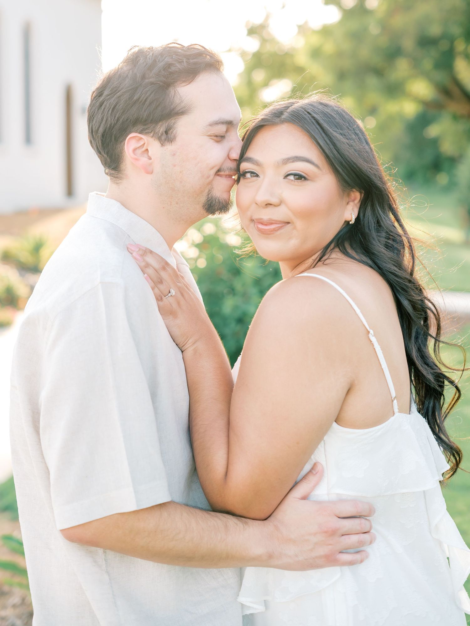 A romantic couple in white outfits embrace outdoors during a sunny engagement photoshoot with natural lighting.