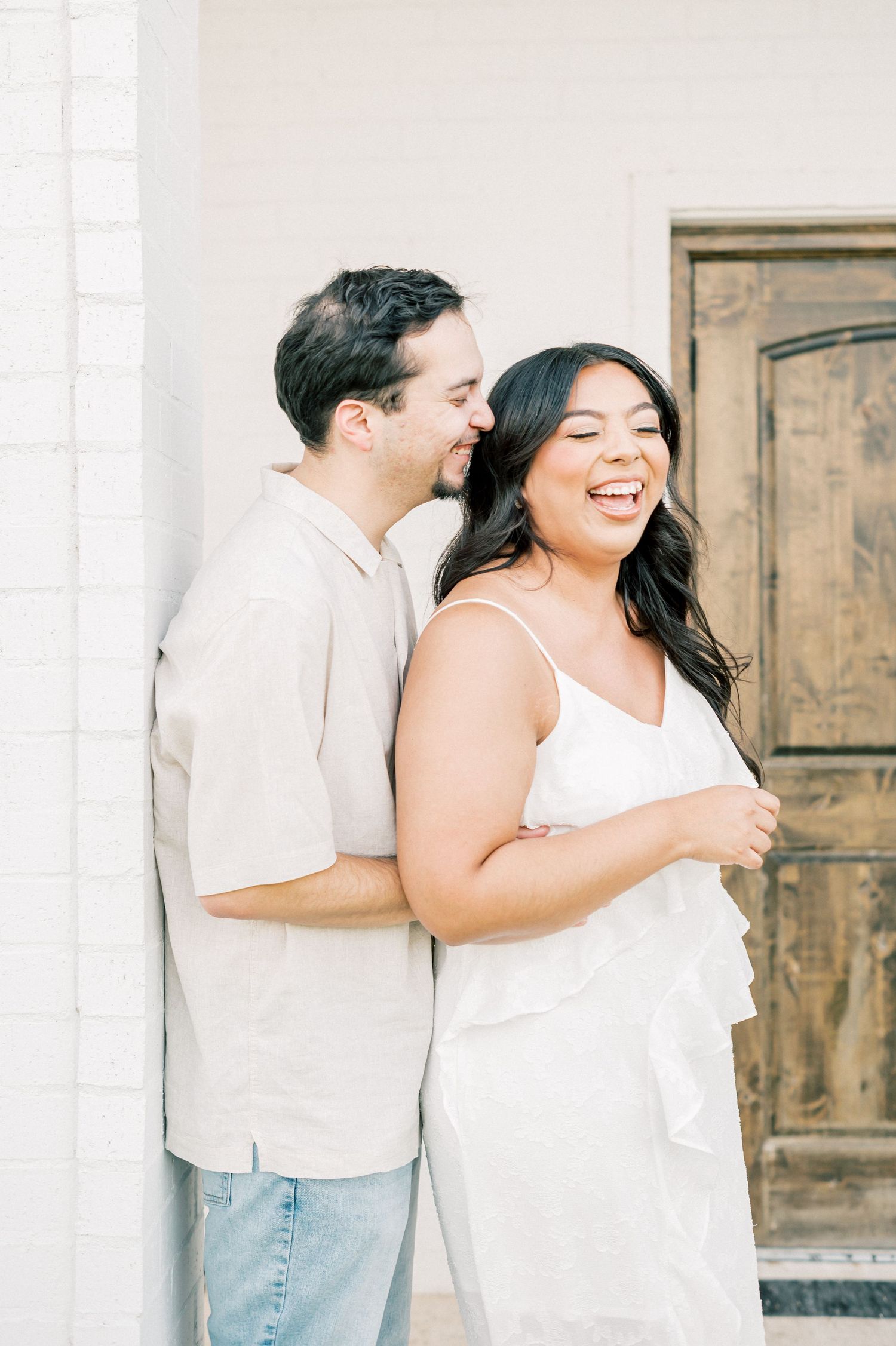 A couple laughing and embracing against a light colored wall during a casual outdoor photo session.
