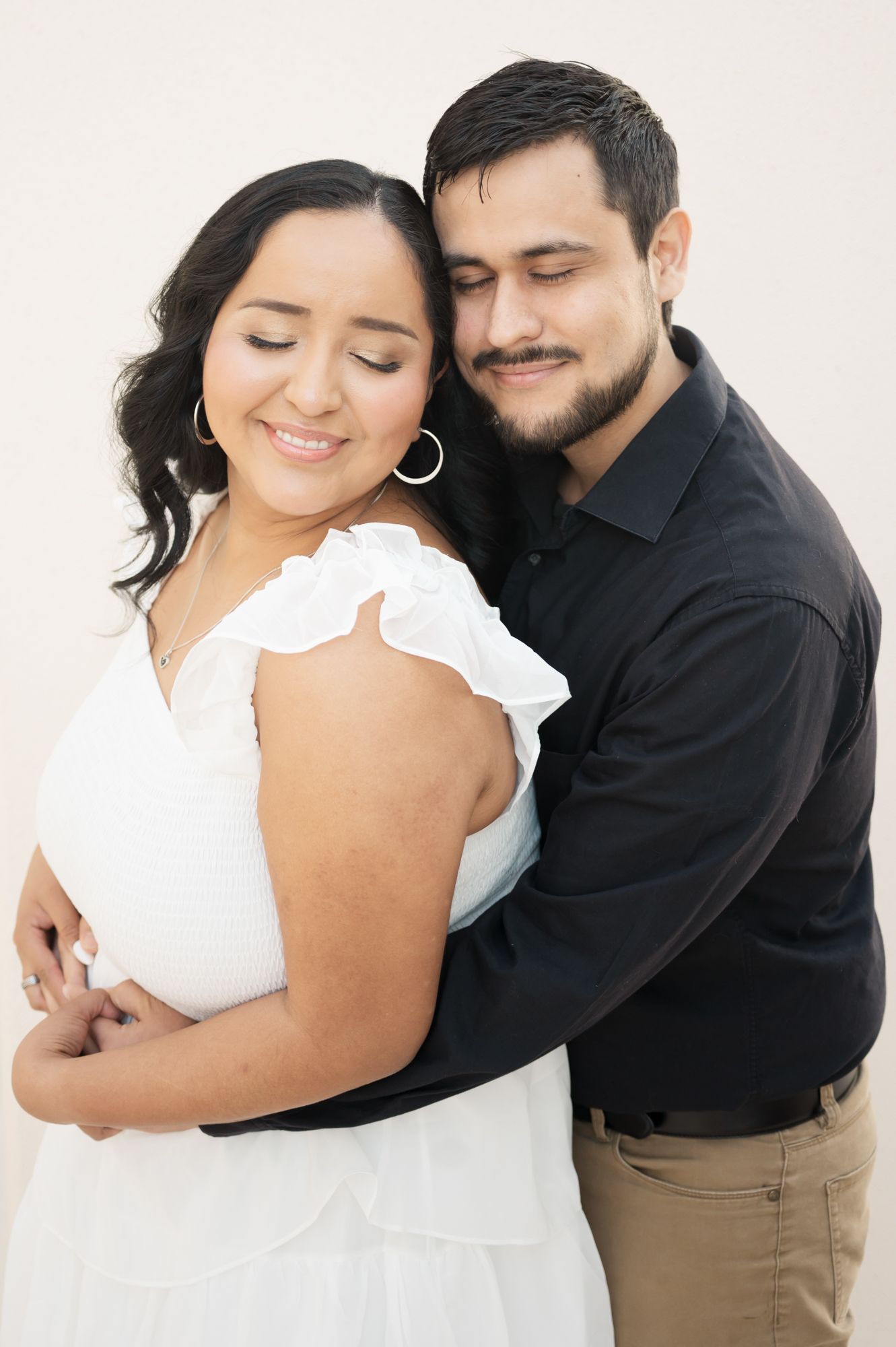 Romantic couple embracing in white outfits against minimal background during engagement photoshoot.