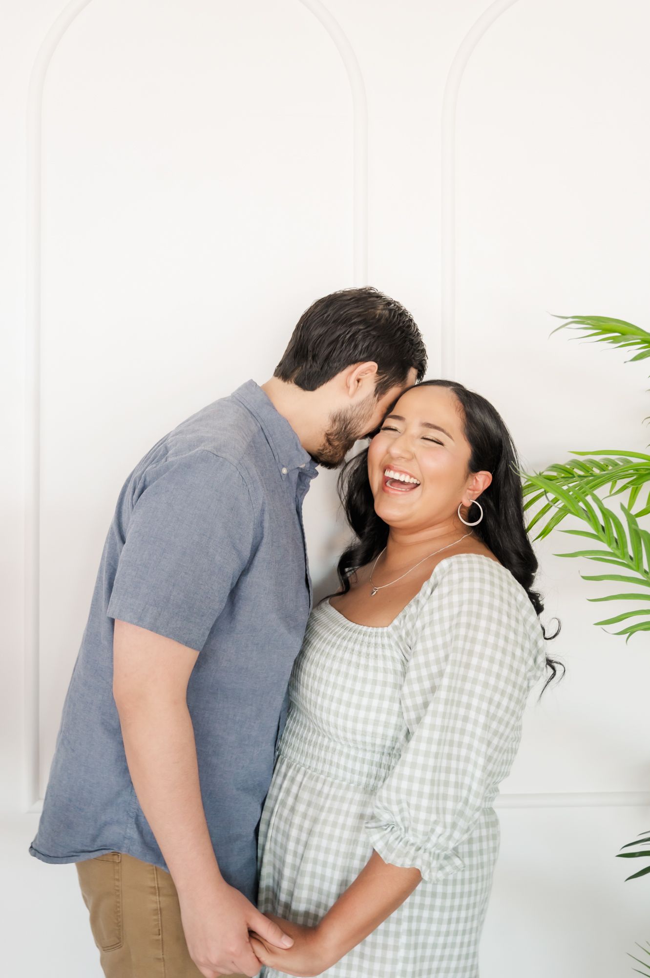 Couple shares intimate moment against white wall with tropical plant in corner.