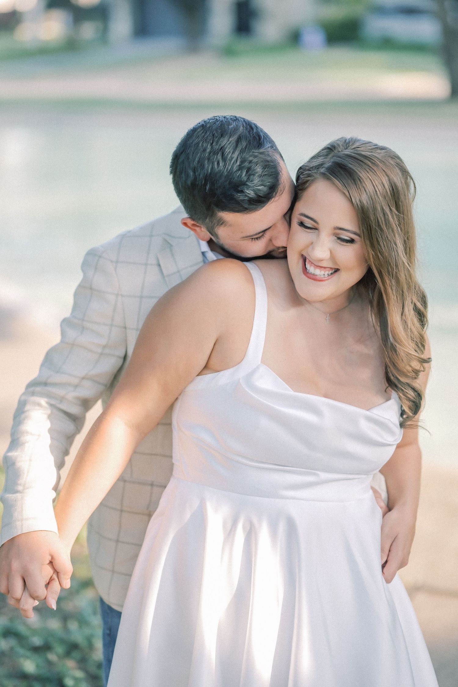 Man in a plaid blazer kisses the cheek of a laughing woman in a white satin dress while they hold hands in a sunny park.