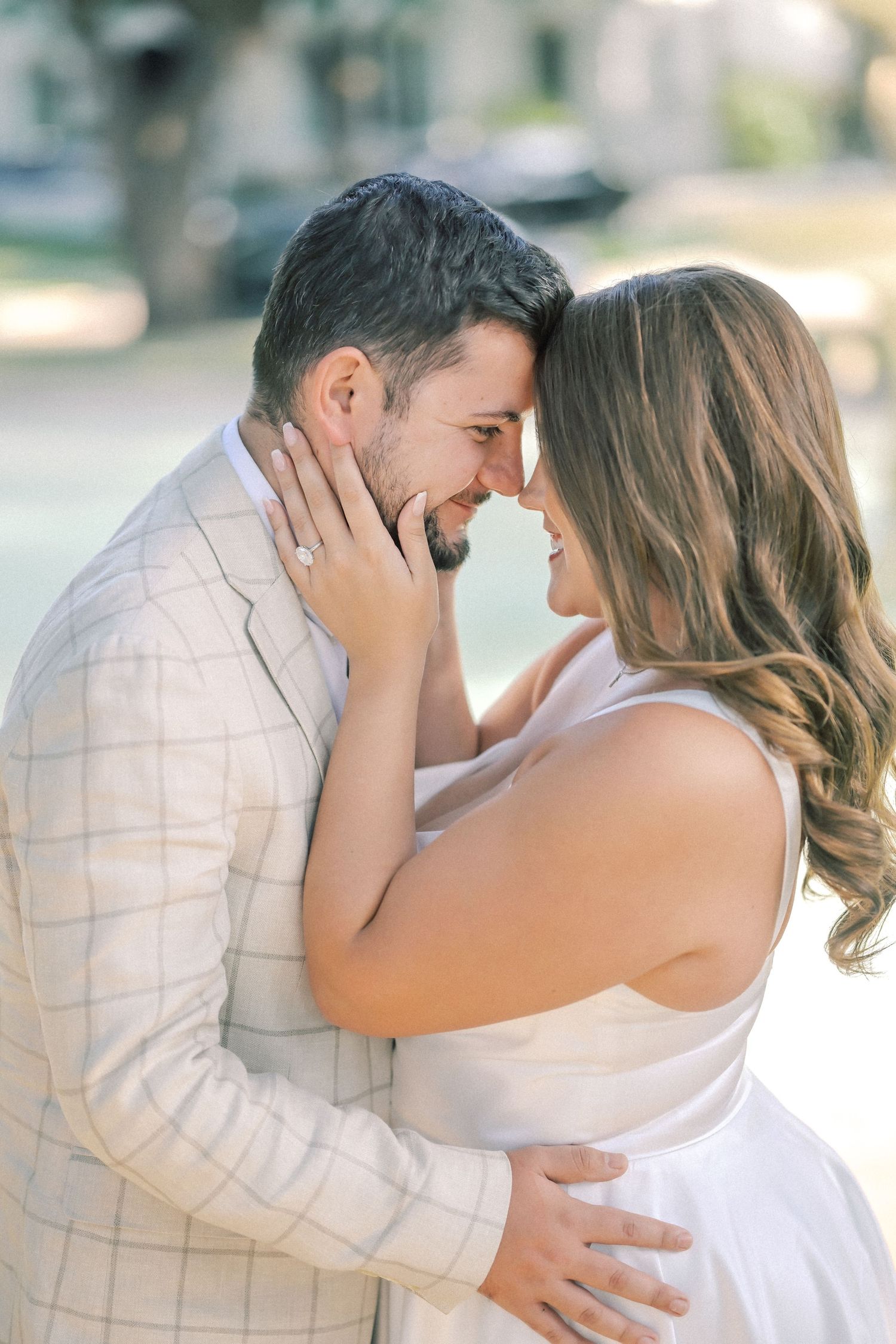 Man in a plaid blazer and woman in a white dress share a forehead touch as she gently cradles his face.