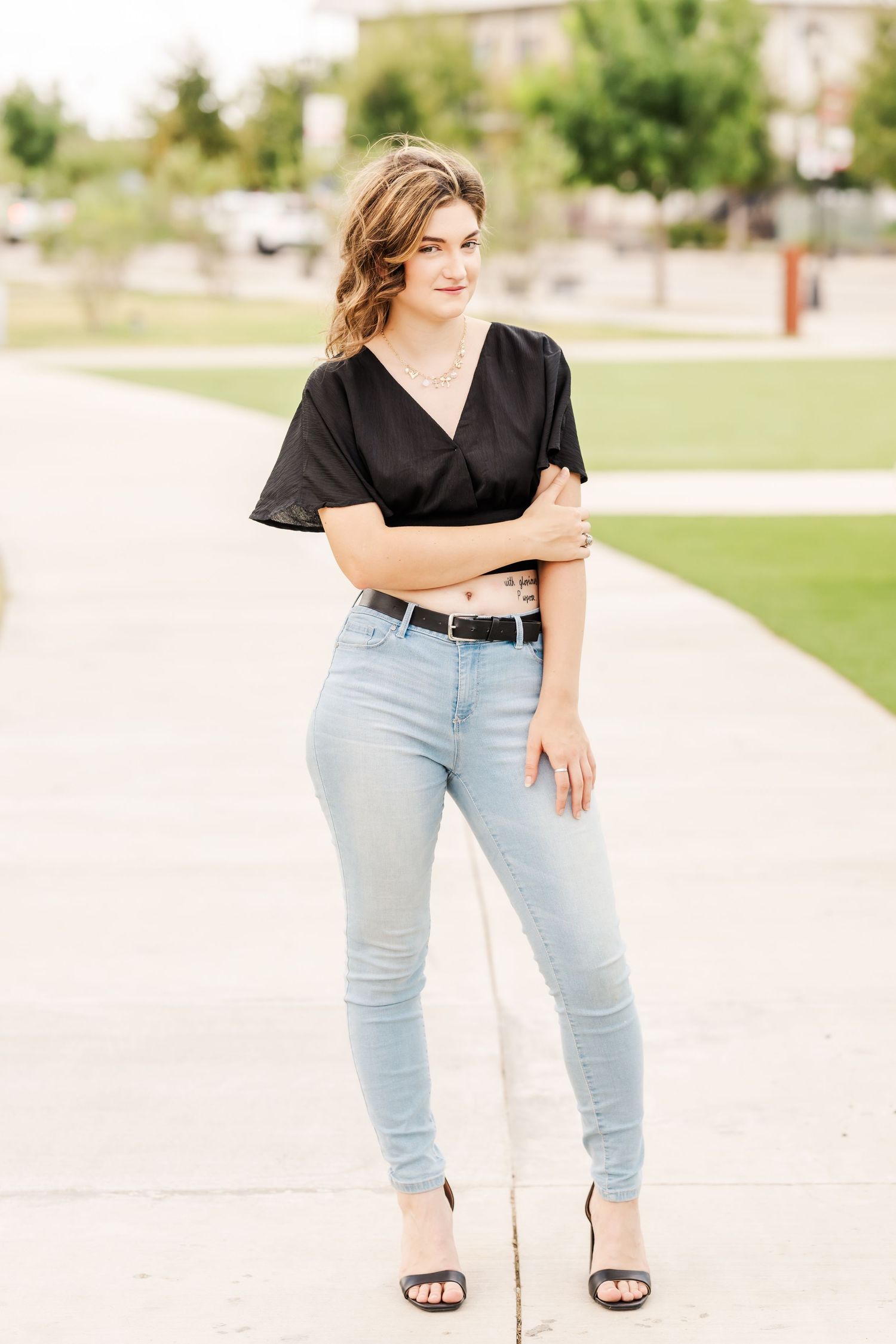 Young woman in black crop top and light wash jeans poses outdoors on a sunny day wearing black heeled sandals.