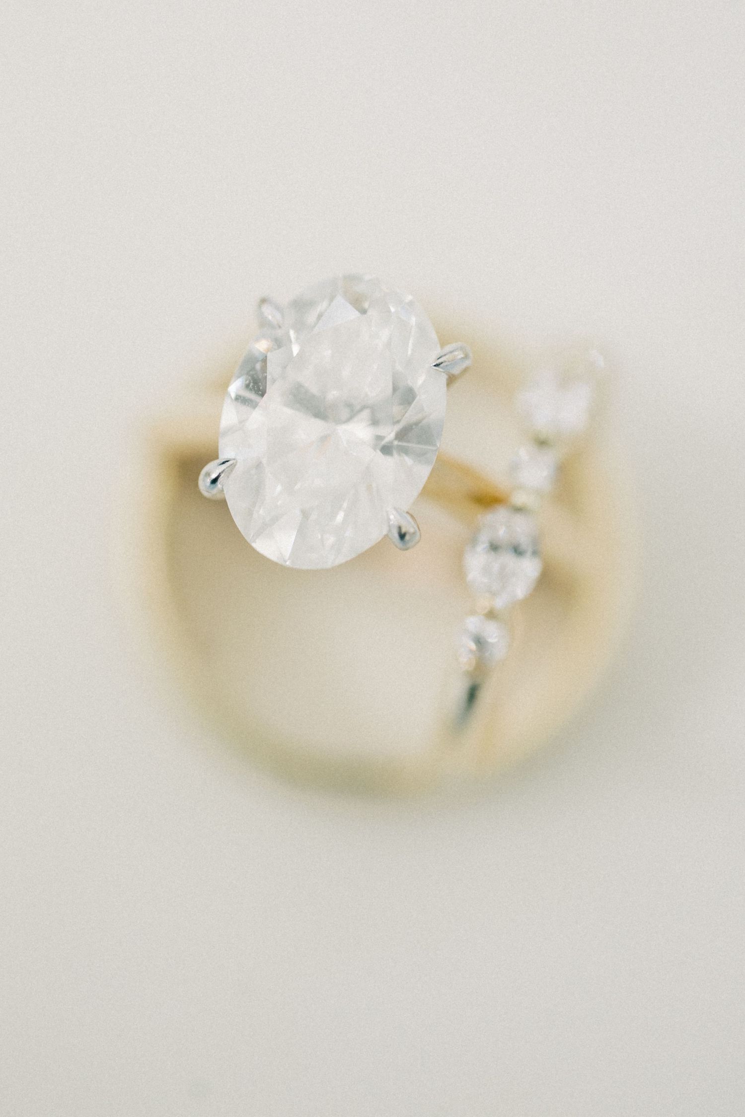 Elegant oval diamond solitaire engagement ring with gold band and accent stones on a soft neutral background.