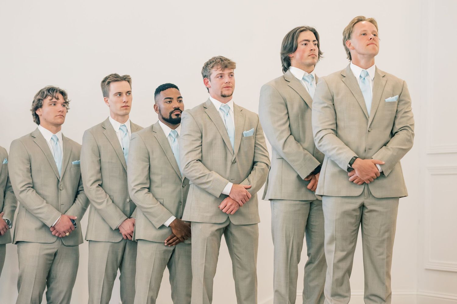 Six groomsmen in tan suits with white ties stand at the altar during a wedding ceremony.