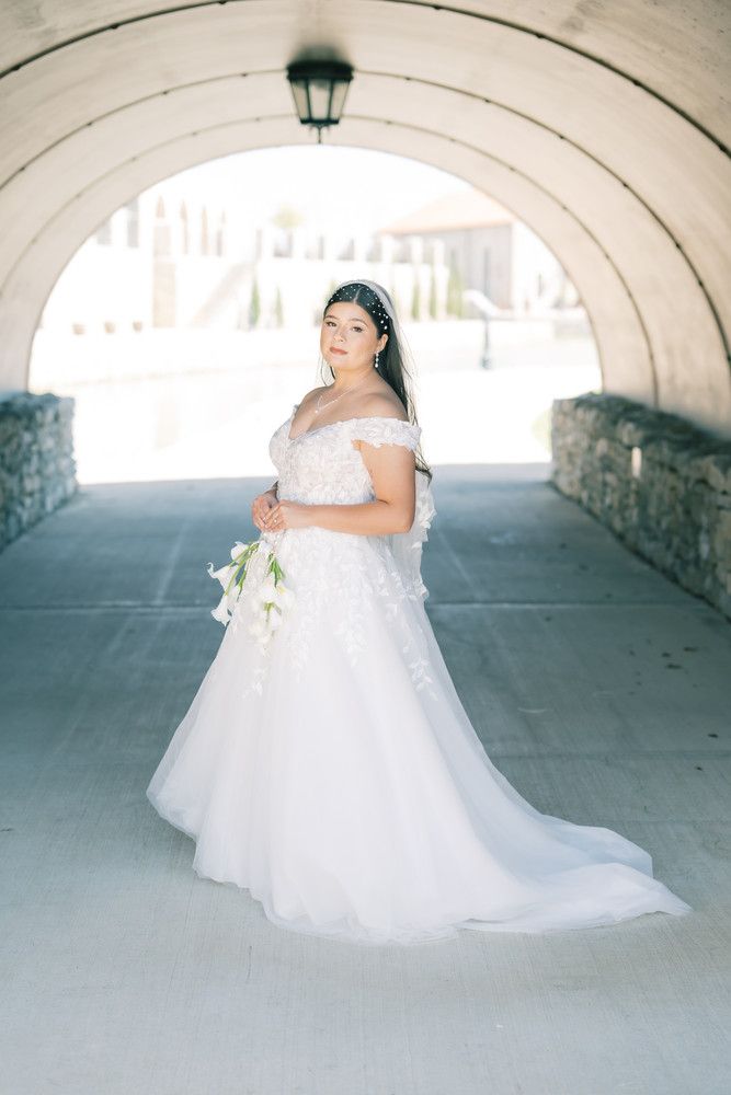 A bride in a flowing white off-shoulder gown twirls gracefully under a stone archway in a romantic bridal portrait series.