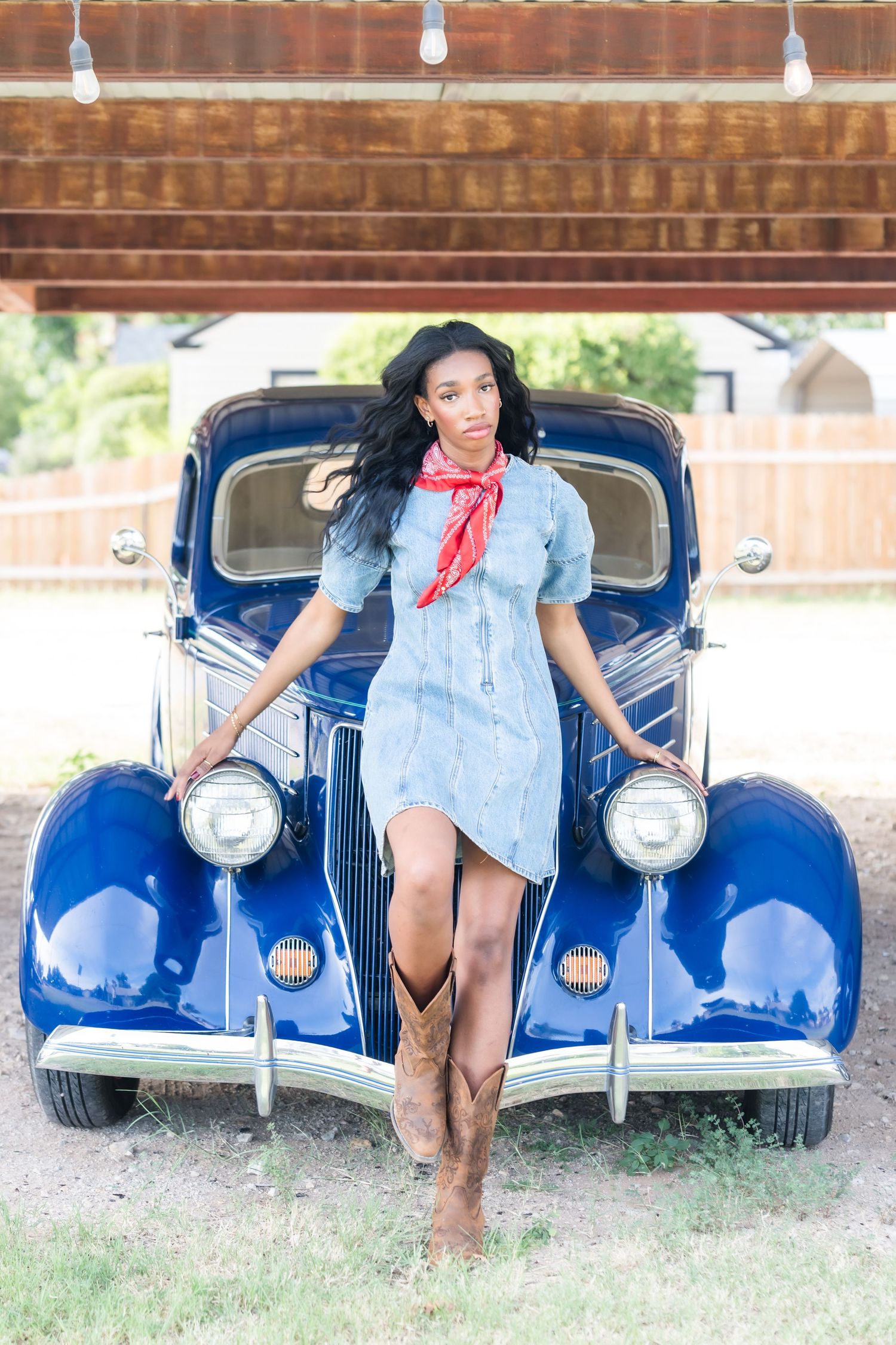 Young woman in denim dress, red bandana, and cowboy boots posing against a classic blue vintage car outdoors.