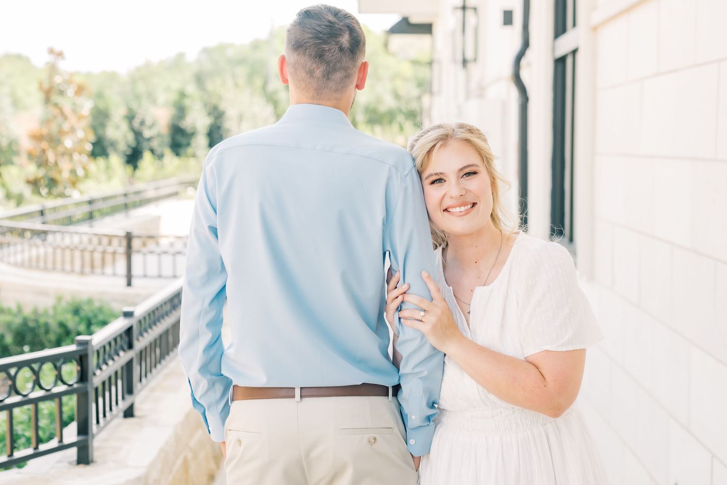 Couple walking on a porch, woman in white dress smiling and showing engagement ring, man in blue shirt.