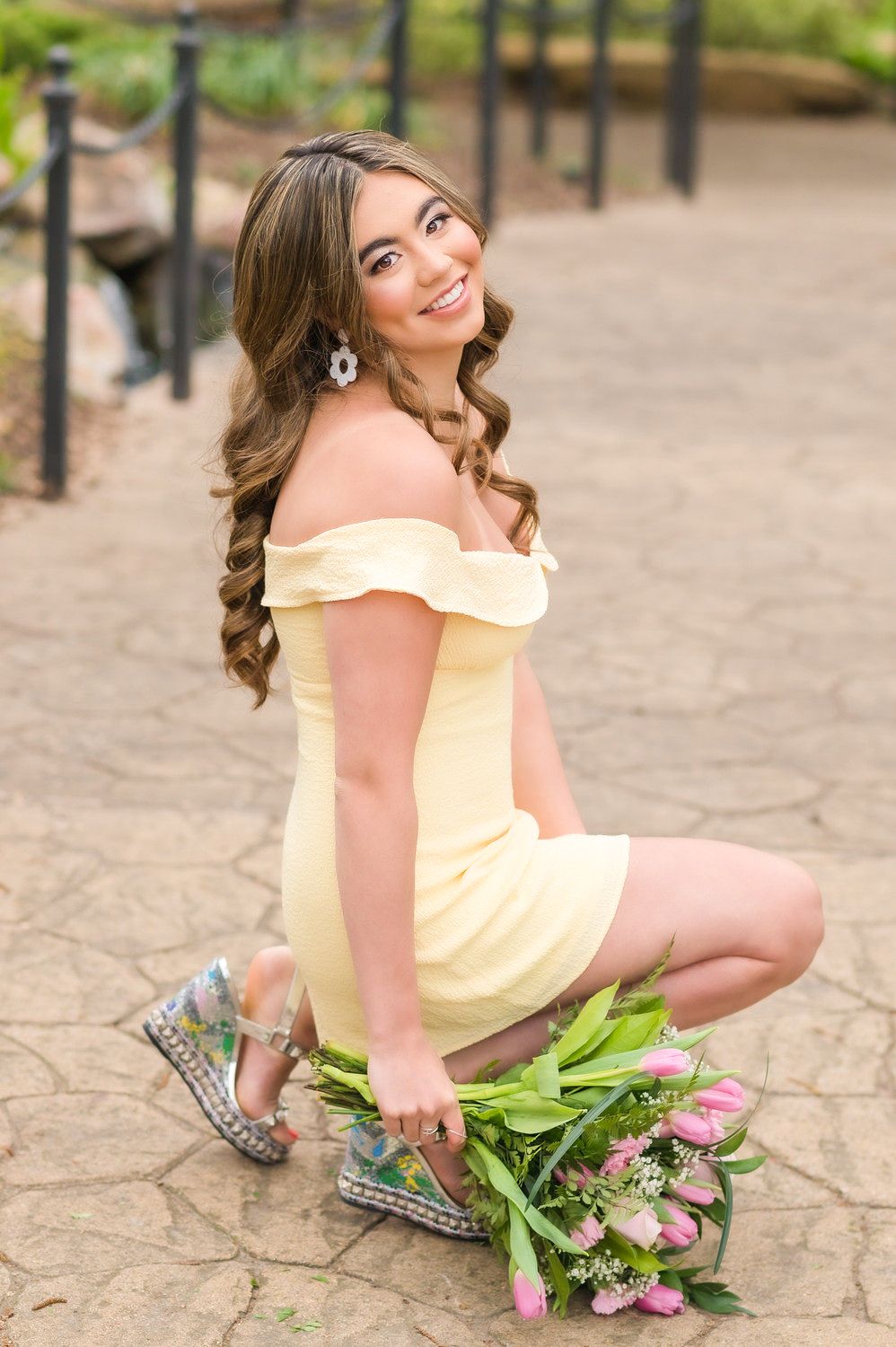Young woman in yellow off-shoulder dress kneeling with pink tulips outdoors.