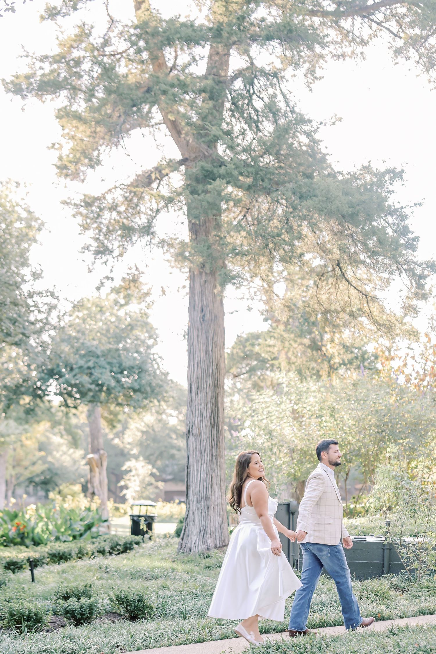 Couple holding hands and walking along a garden path beneath tall trees, she in a white dress and he in a beige jacket and jeans.